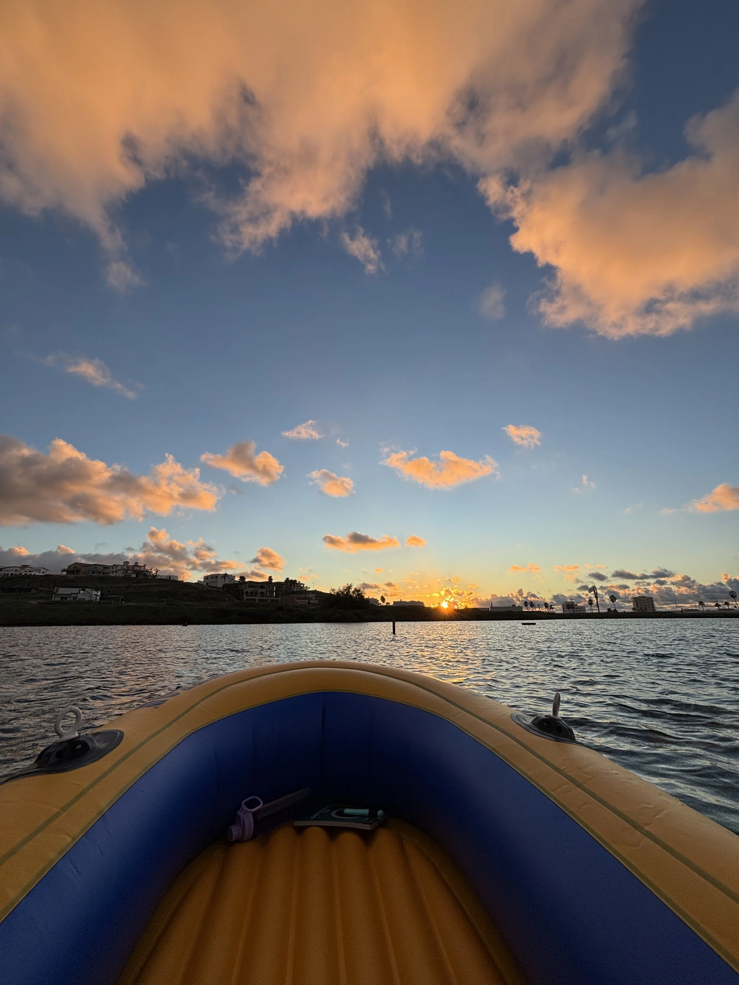 View from a boat on water at sunset with clouds and distant land with buildings and palm trees.