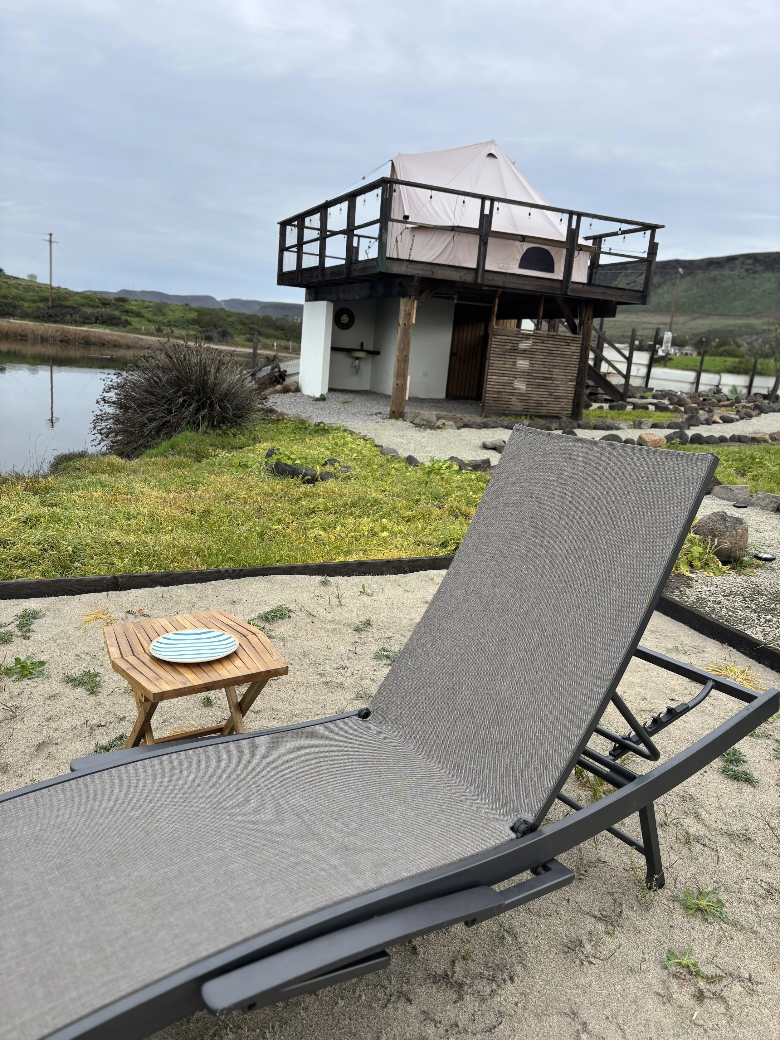 A beach scene with a beige lounge chair and a small wooden table with a striped plate, near a pond. In the background, a raised hut with a tent-like top and wooden stairs is visible, surrounded by rocks, grass, and a landscape of hills and cloudy sky