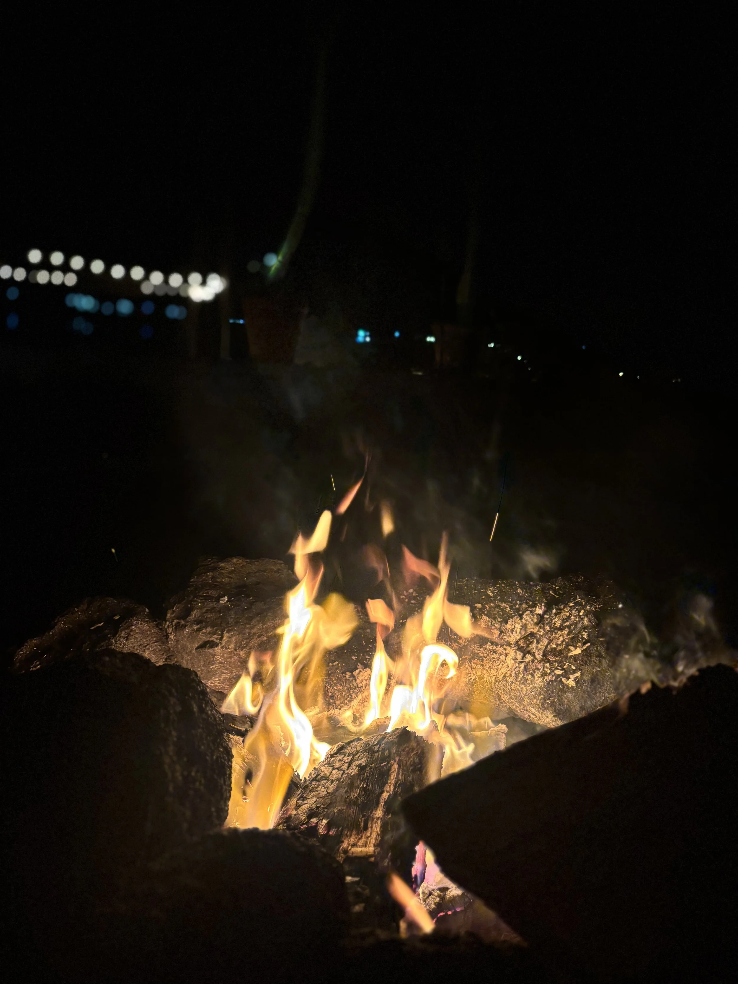 A close-up of a campfire with flames burning among rocks at night, with blurry city or street lights in the background.