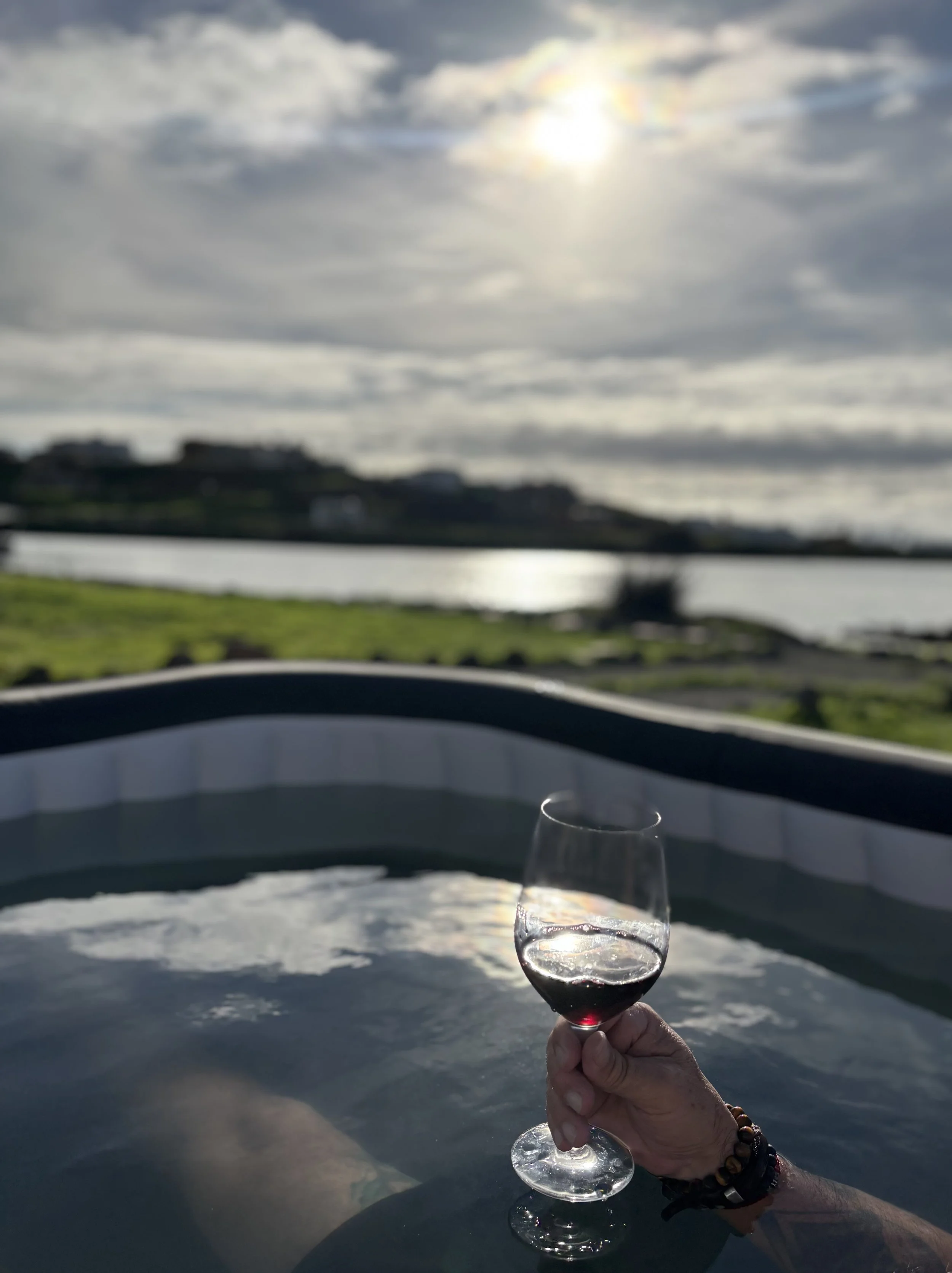Person holding a glass of red wine in a hot tub outdoors during sunset, with a view of a lake and cloudy sky.
