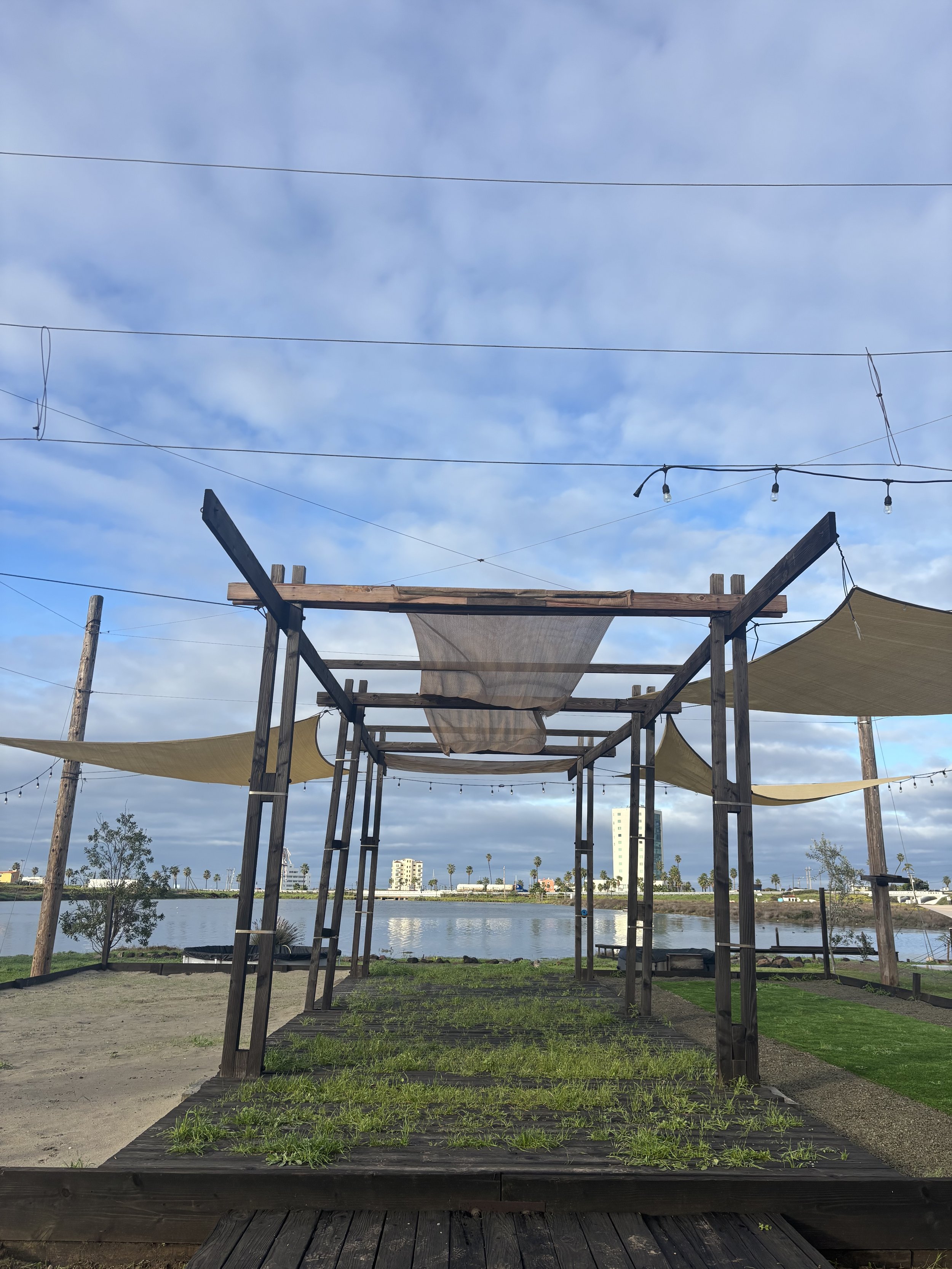 A lakeside outdoor structure with a wooden frame and fabric canopies, set on a grassy and sandy area, with city buildings and palm trees in the background under a cloudy sky.