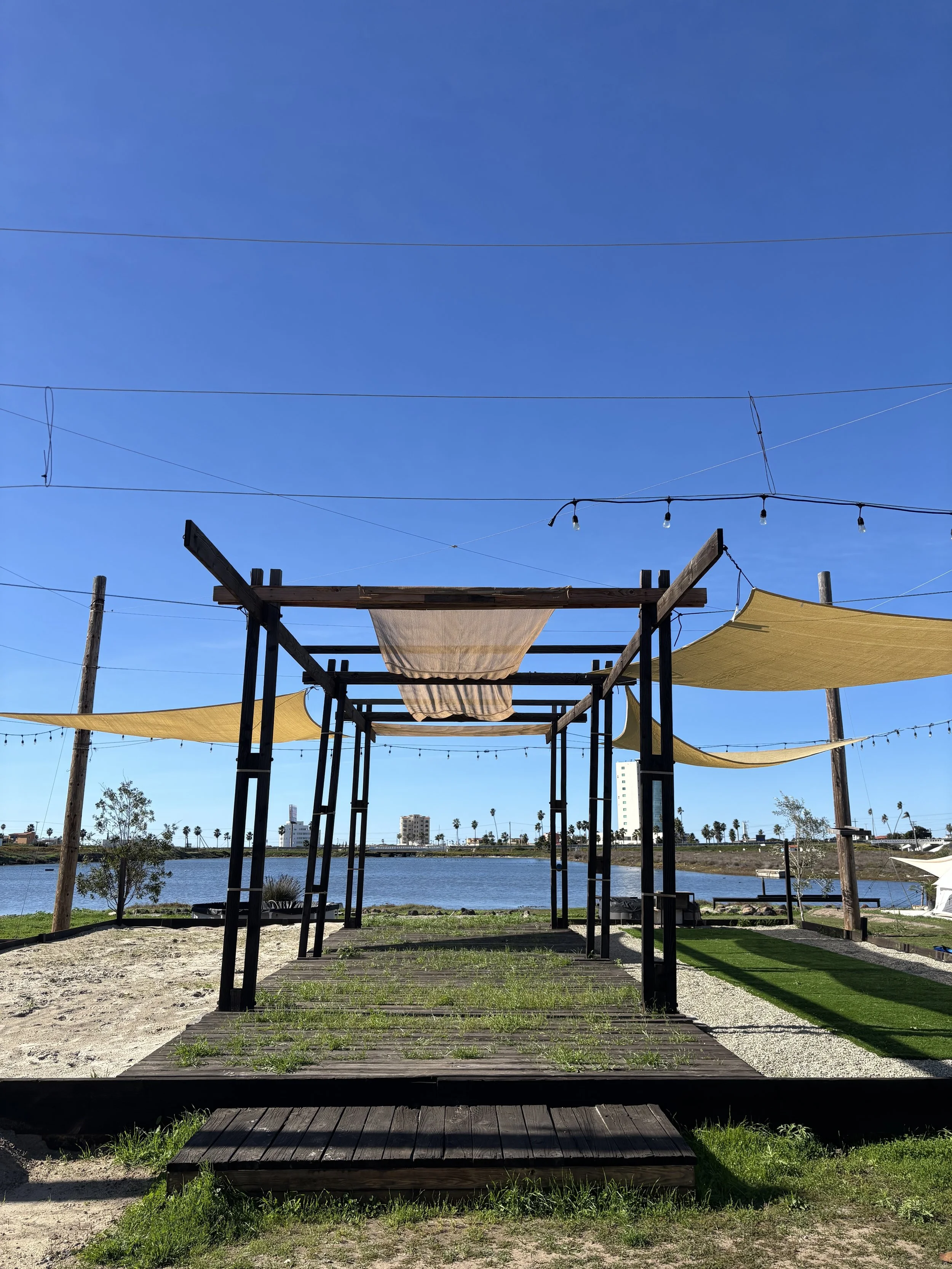 Outdoor patio with wooden deck, black pergola, yellow shade sails, and string lights overlooking a body of water and city skyline in the background.