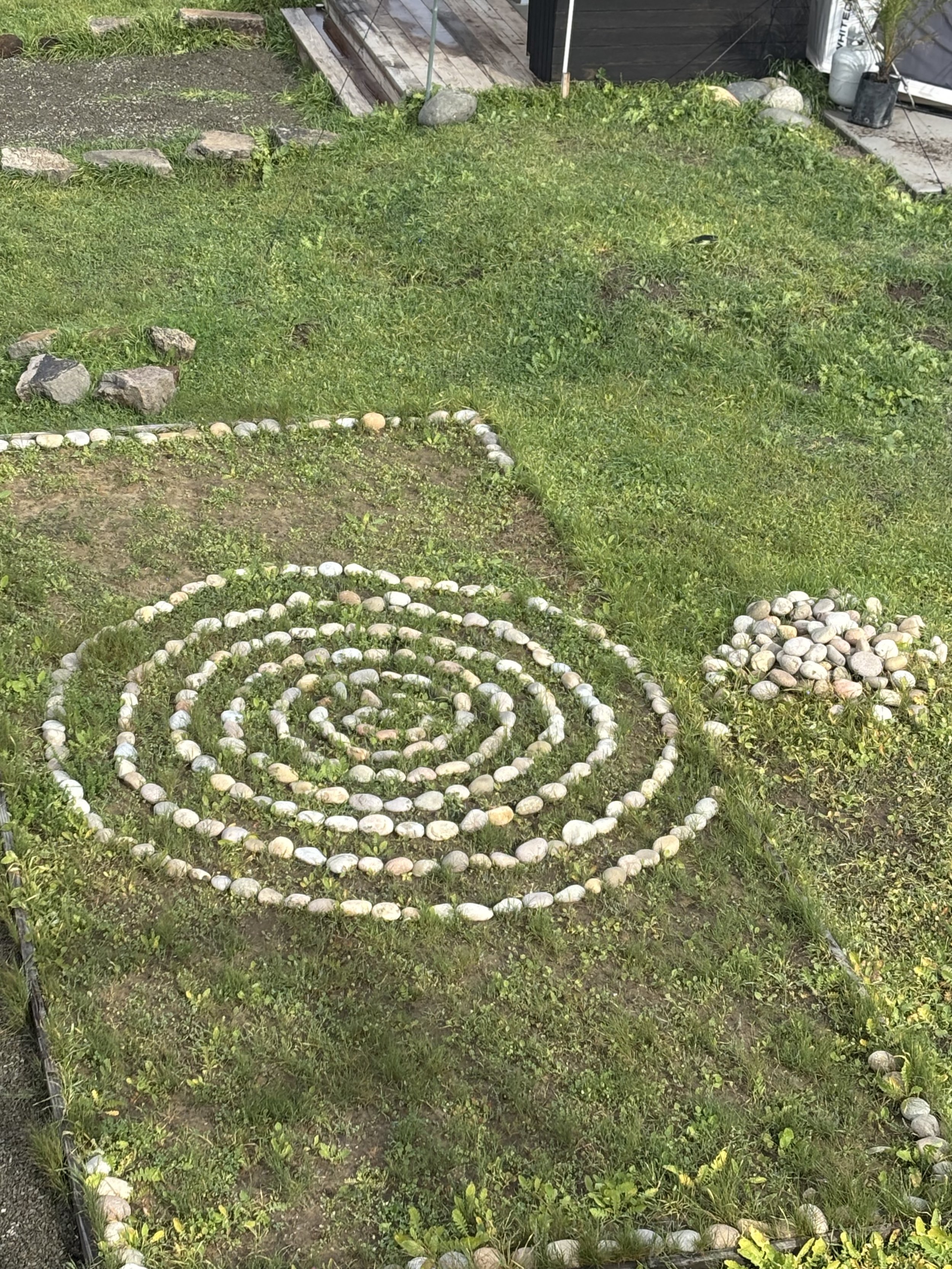 A garden with spiral-shaped arrangements of rocks and stones on grass, with additional rocks and a black wooden structure in the background.