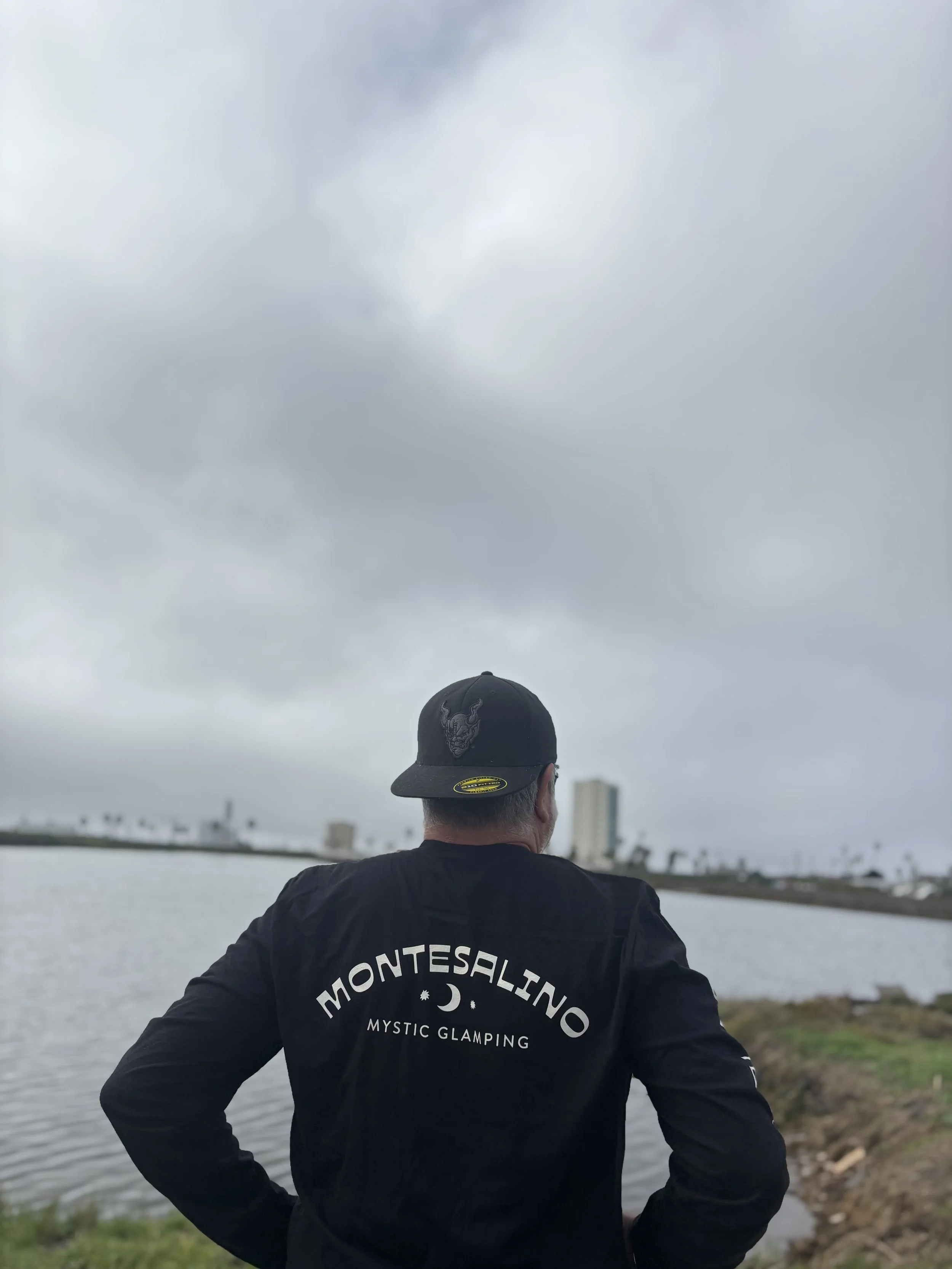 Man wearing a black MontIsAlino Mystic Glamping jacket and a black cap with a dragon emblem standing by a body of water on a cloudy day, with buildings and palm trees in the distance.