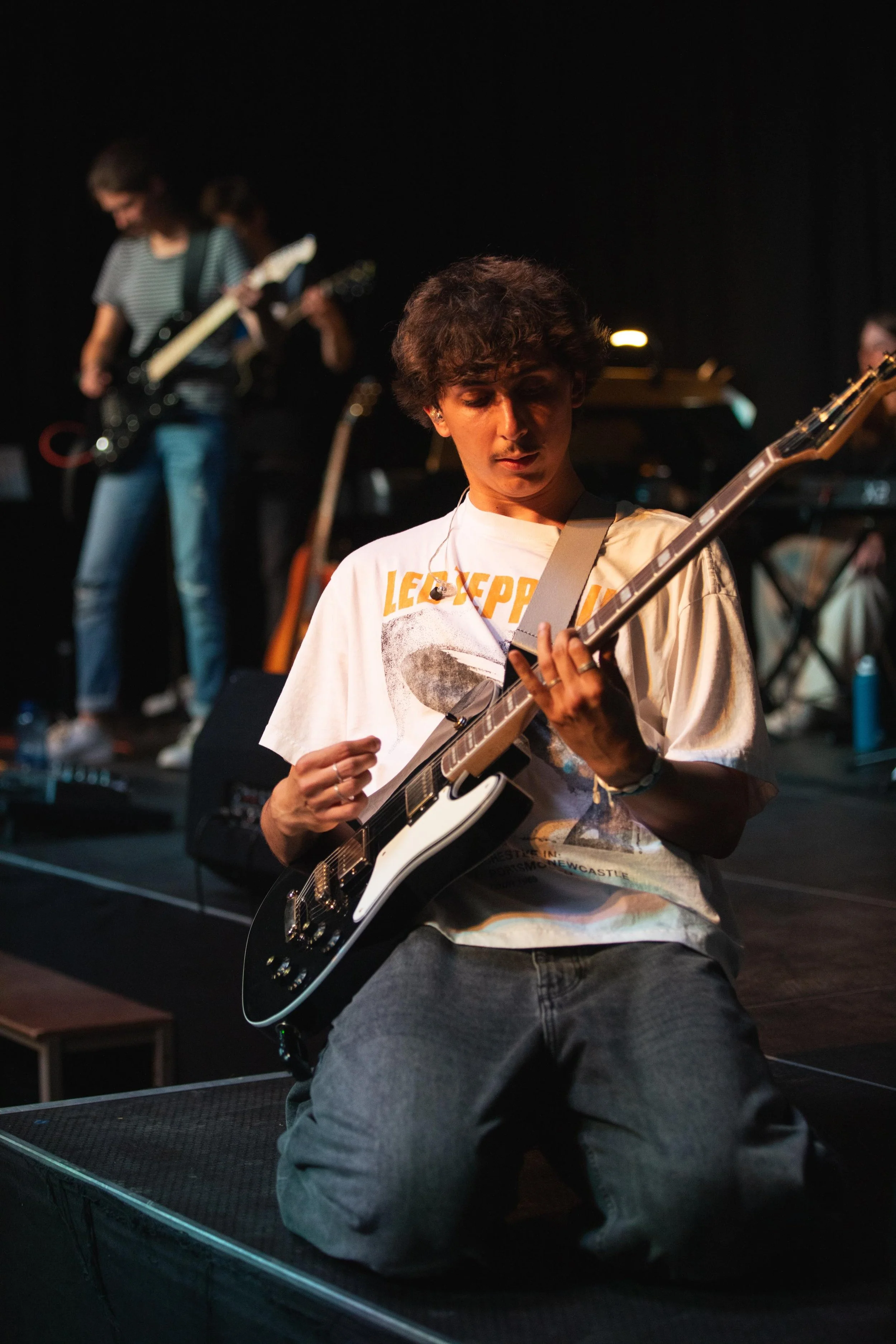 Eine junge Person, die auf Knien auf der Bühne sitzt und Gitarre spielt, während im Hintergrund andere Bandmitglieder spielen.