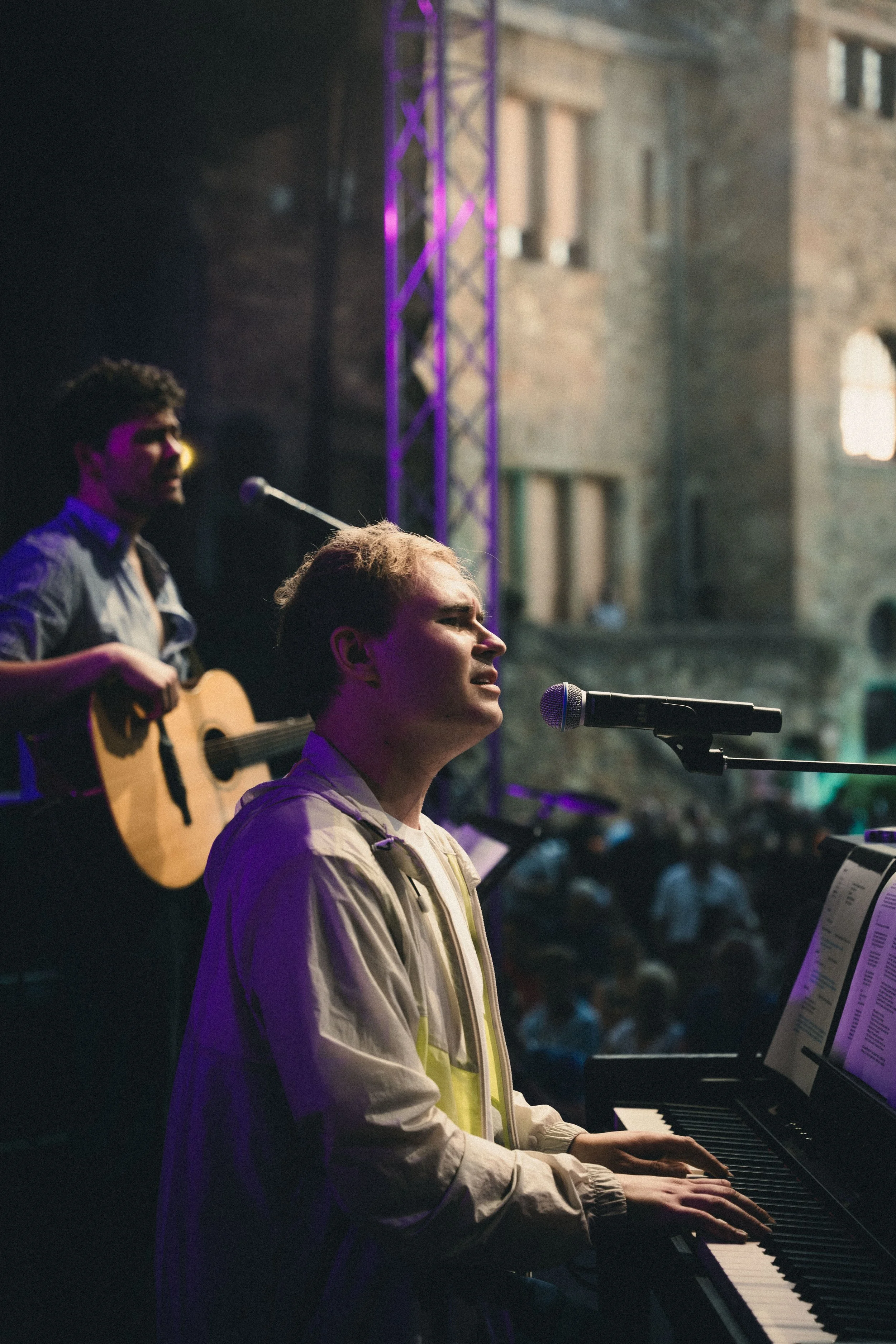 Zwei Musiker bei einem Live-Konzert im Freien, einer spielt Klavier und singt, der andere spielt Gitarre, im Hintergrund ein altes Gebäude und Publikum.