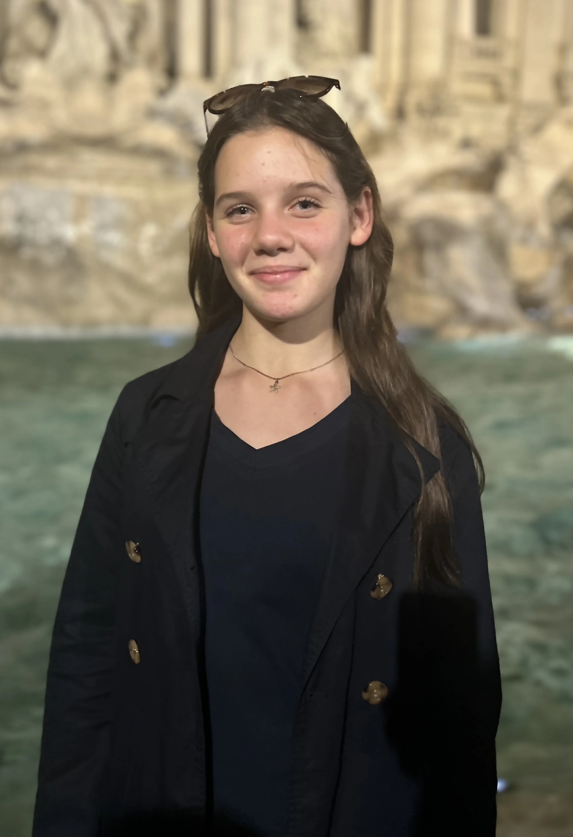 Junge Frau mit braunen Haaren und Sonnenbrille auf dem Kopf, trägt eine schwarze Jacke und ein schwarzes T-Shirt, steht vor einer Felswand und Wasser.