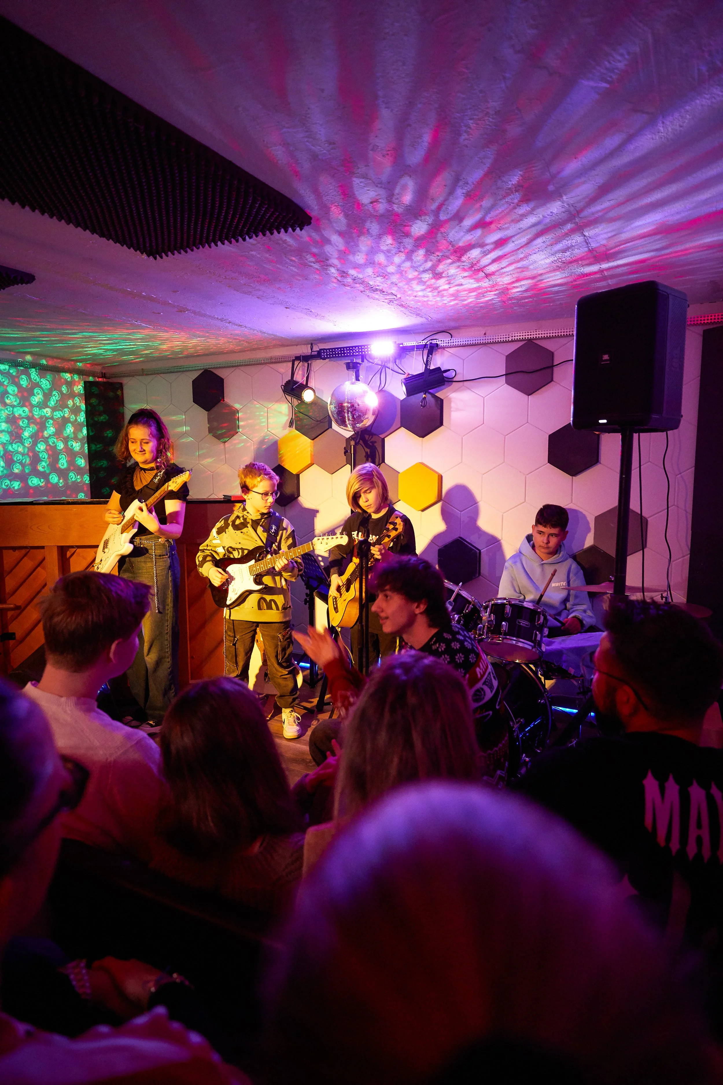 Jugendliche spielen Gitarre und Schlagzeug auf einer kleinen Bühne vor Publikum in einem Raum mit buntem Licht und Wanddekorationen aus hexagonalen Mustern.