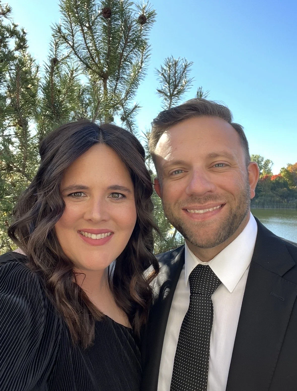 A smiling man and woman taking a selfie outdoors by a lake with trees and a blue sky in the background.