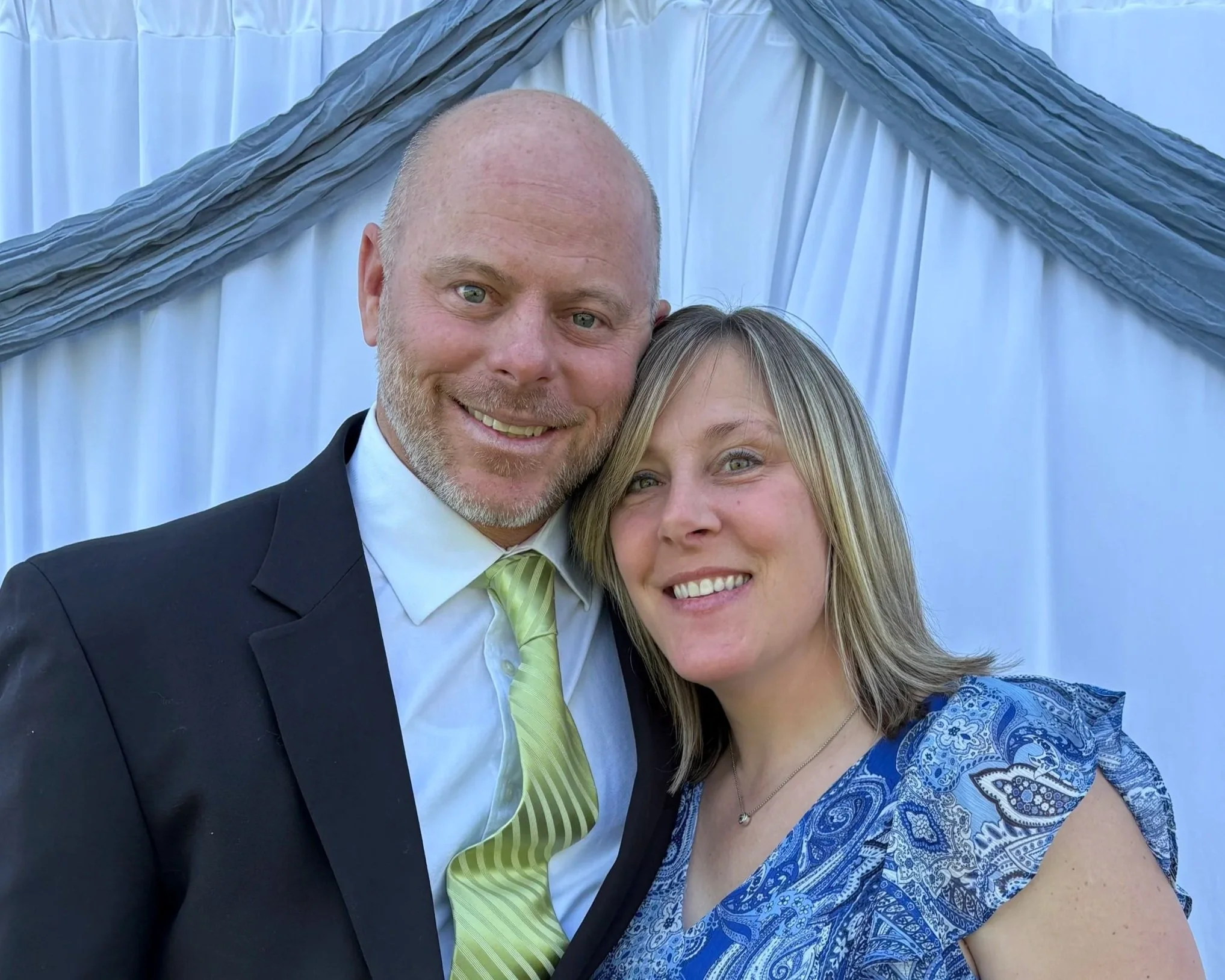 A smiling man and woman posing closely together at an outdoor event. The man is dressed in a black suit with a white shirt and a green striped tie. The woman is wearing a blue patterned dress with ruffled sleeves and a small pendant necklace. Behind them is a blue and gray draped fabric backdrop.