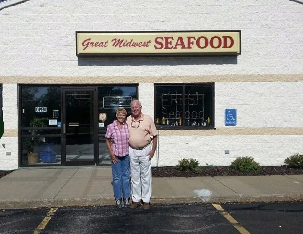 Two people standing in front of a restaurant with a sign that reads "Great Midwest Seafood." The restaurant has a large window and a door with an "Open" sign, and a handicap parking sign is visible nearby.