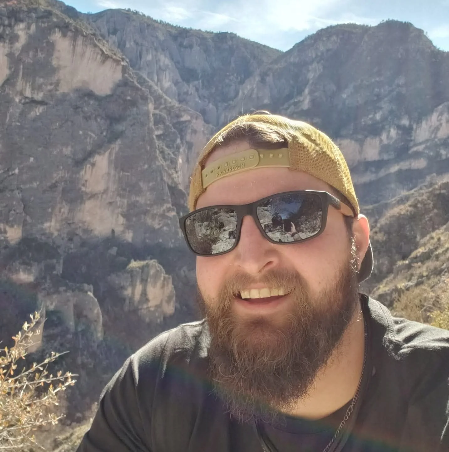 A man taking a selfie outdoors in front of a mountain canyon, wearing sunglasses, a yellow cap backwards, and a black shirt.