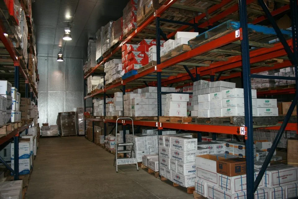Warehouse aisle with metal shelving filled with stacked boxes, some labeled with logo and text, and a small ladder in the center.