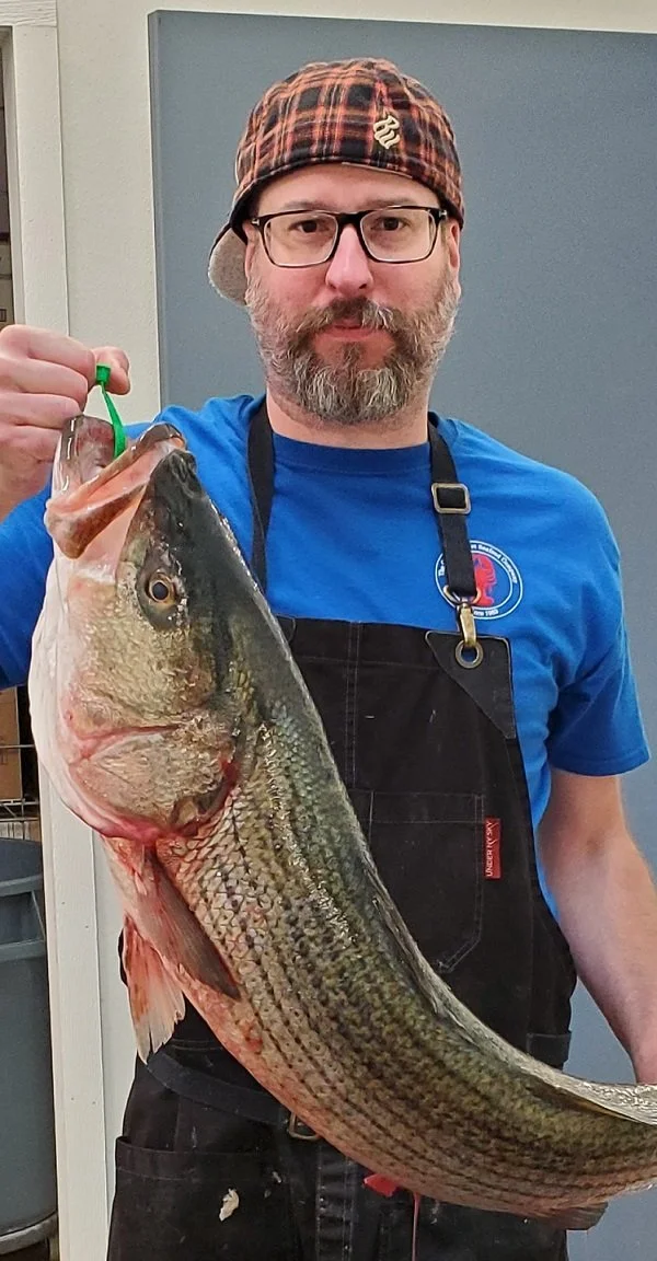 Man with glasses, beard, and plaid cap holding a large fish, possibly a bass, indoors with gray and white walls.