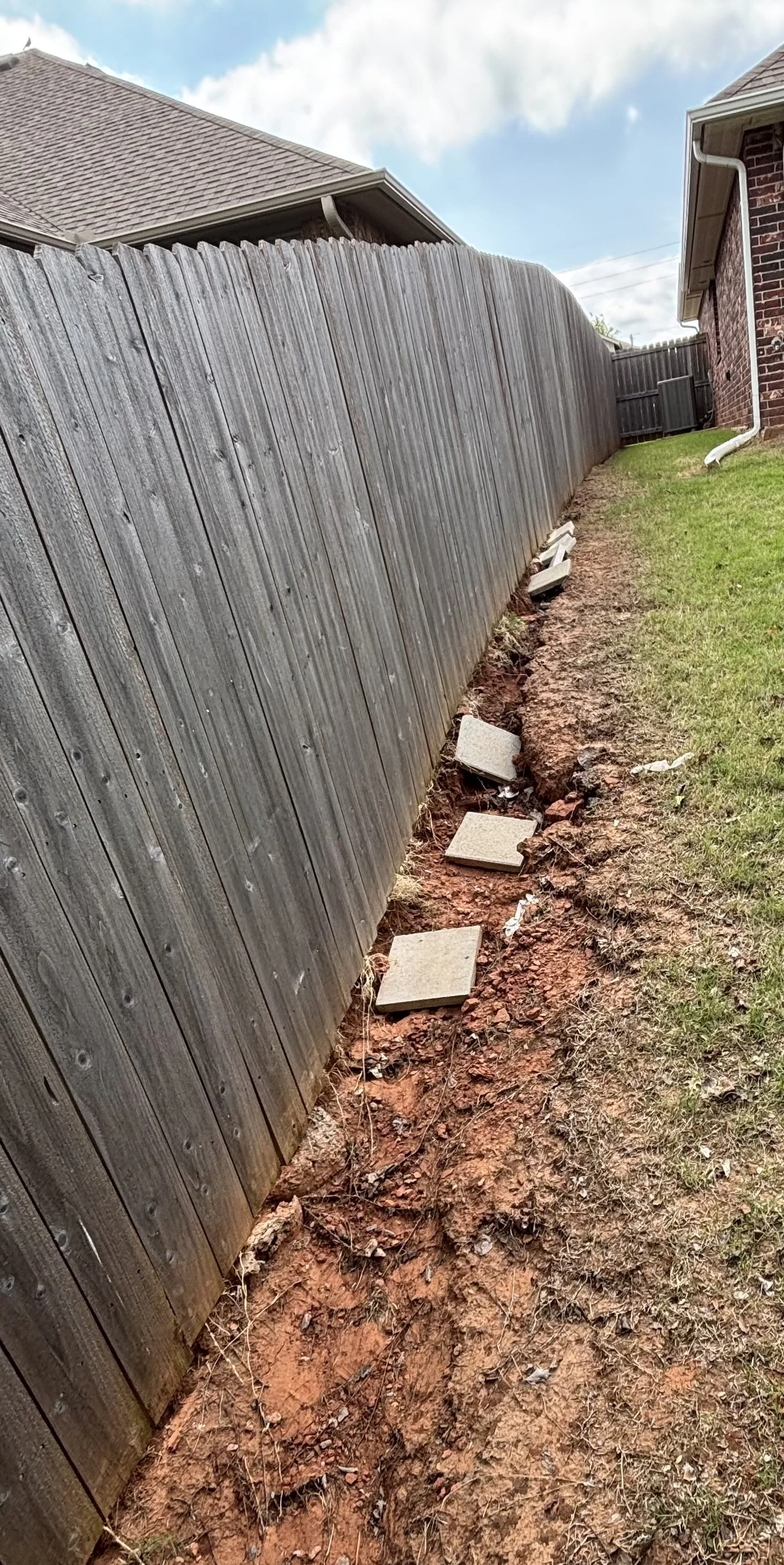 A wooden fence with some missing and replaced concrete stepping stones on the ground along the fence line, with a brick house on the right side and partly cloudy sky overhead.