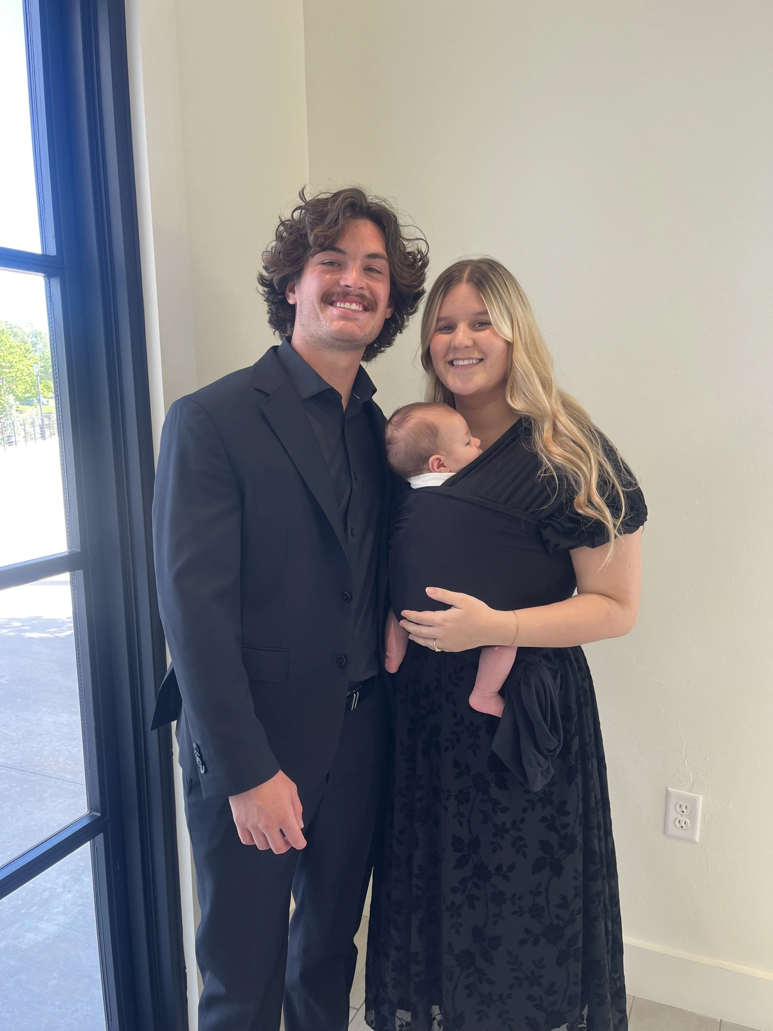 A man in a black suit and a woman in a black dress holding a baby. They are standing next to a blue-framed door with a window, inside a room with light-colored walls and an electrical outlet visible.