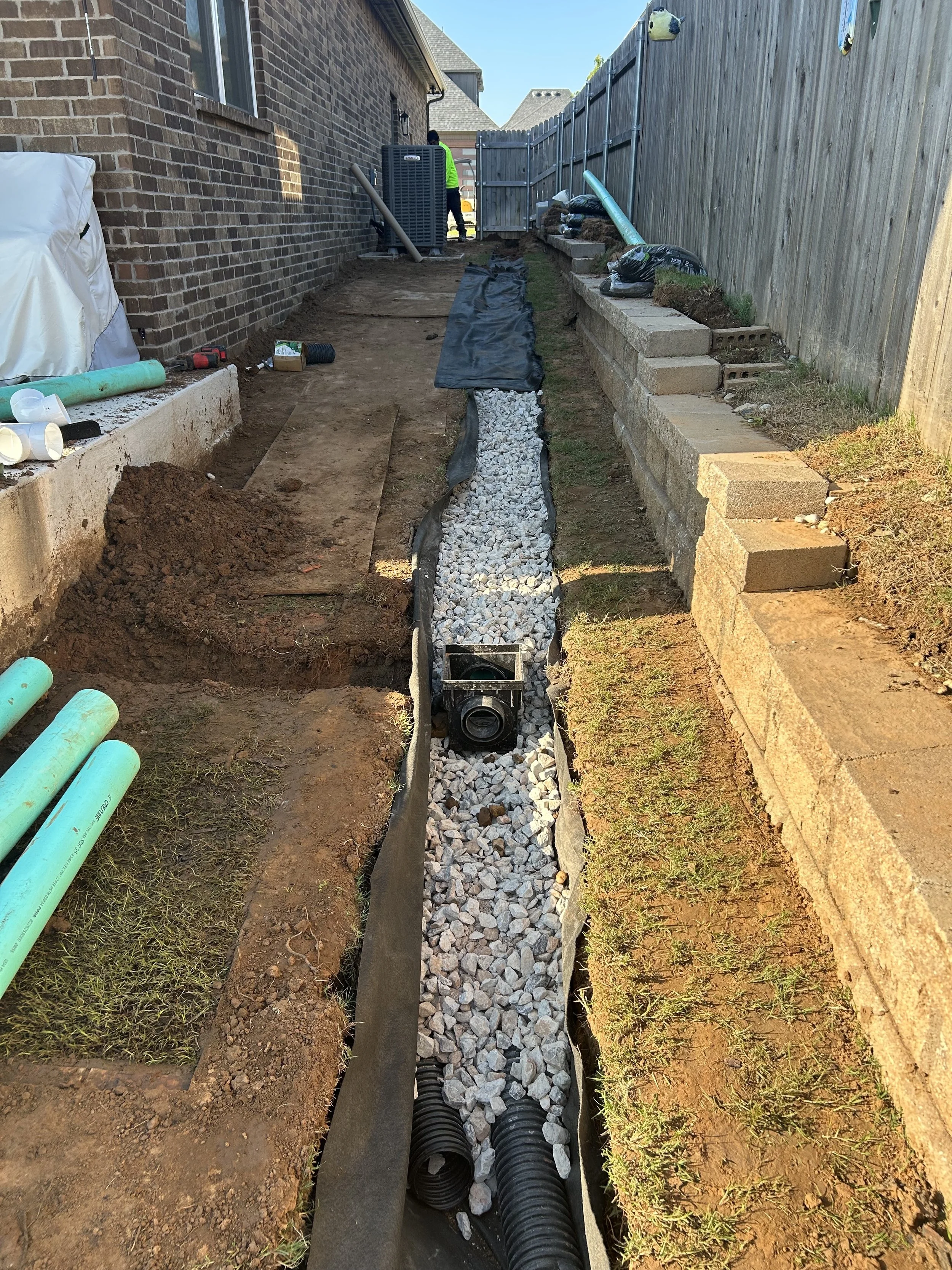 A backyard under construction with a gravel trench filled with white rocks, black corrugated piping, and construction tools. There is a brick house on the left and a wooden fence on the right. Construction materials and equipment are scattered around.