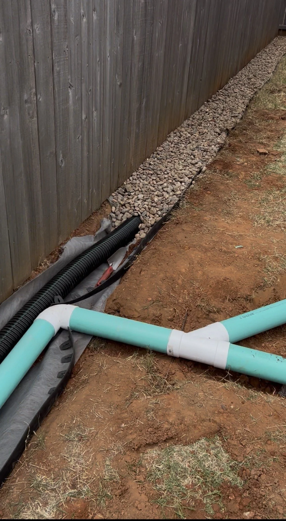 Underground piping and drainage system installation next to a wooden fence, with gravel and soil.