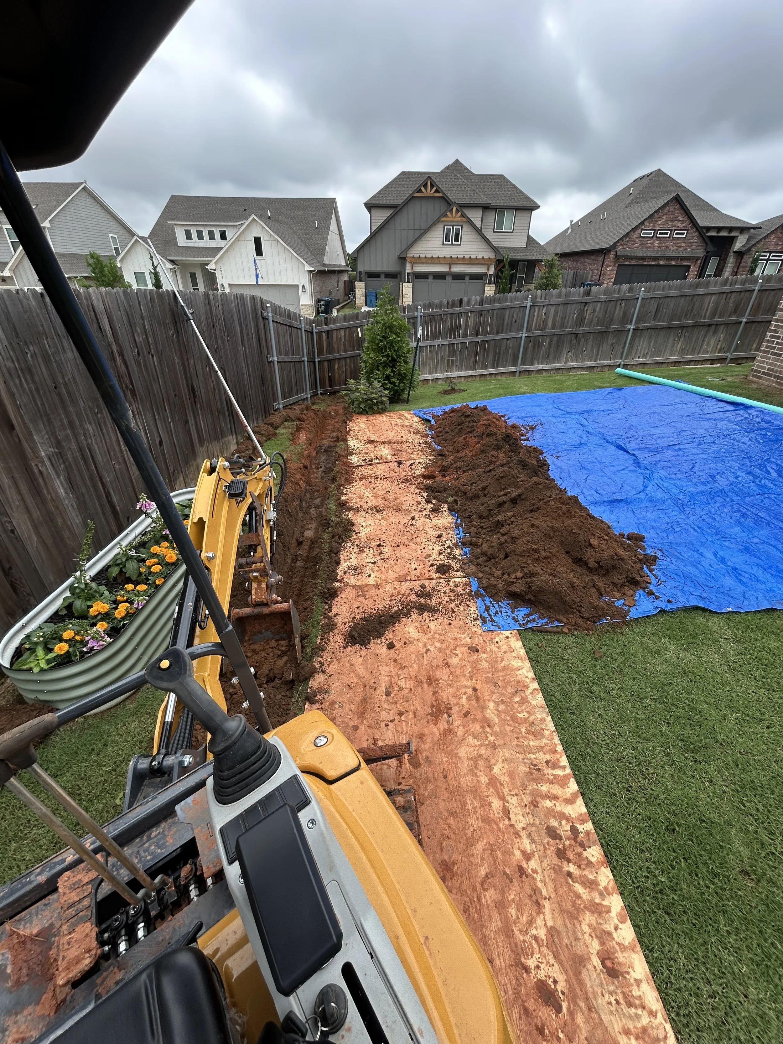 A backyard scene showing a wooden fence, flower beds, and a yard covered with a blue tarp and dirt, with construction equipment and tools visible in the foreground.