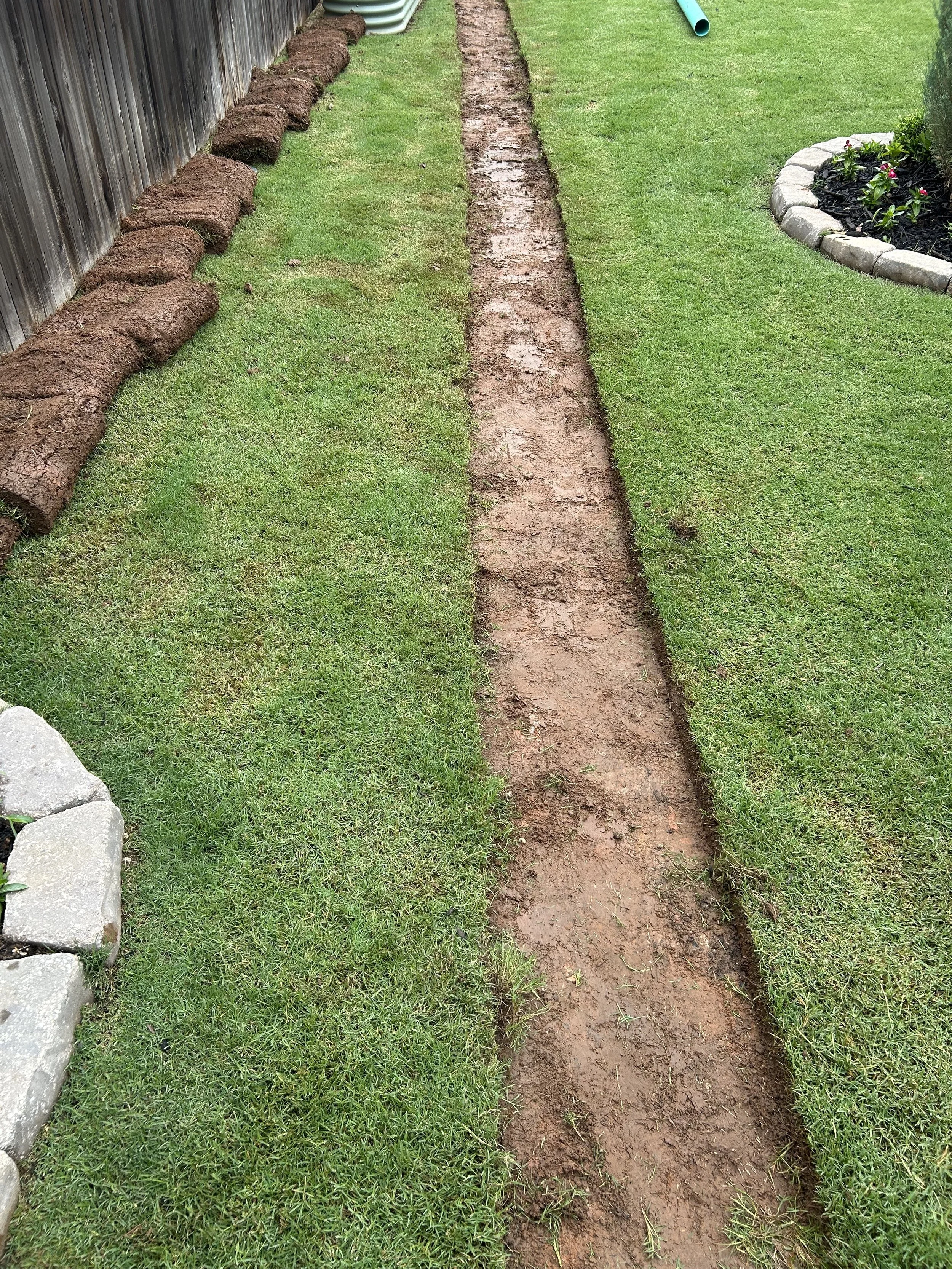 A garden with a narrow dirt pathway running through the grass, bordered by a wooden fence on the left and a flower bed with rocks on the right.