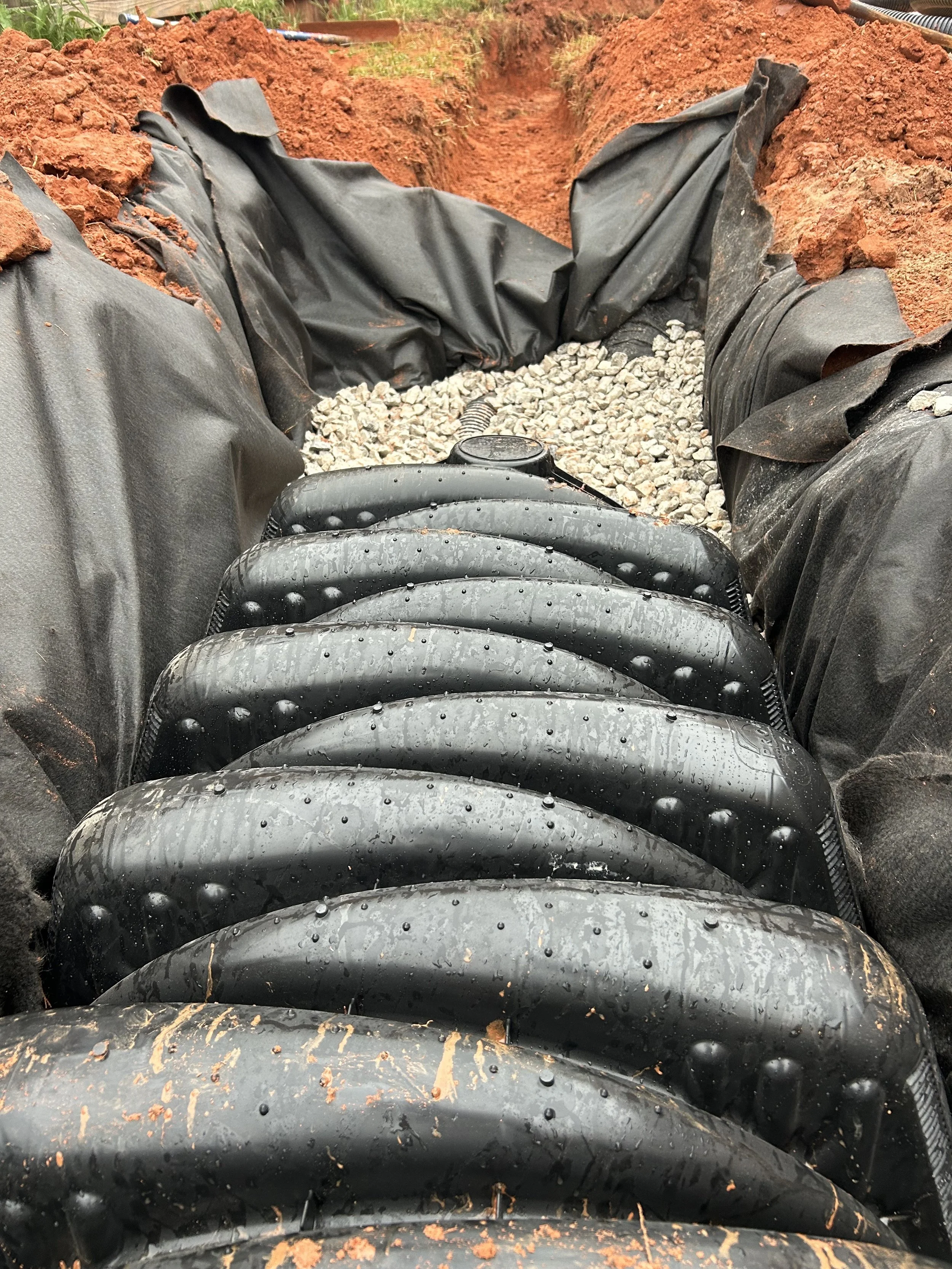 Close-up of black pond liners and a drainage pipe laid out in a trench, with dirt and gravel visible, likely as part of a pond or water feature installation.
