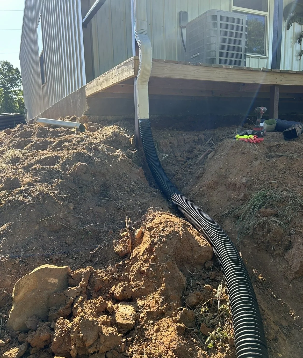 A black corrugated drainage pipe runs through a dug trench in dirt, leading towards the foundation of a house with a raised wooden deck. Construction tools, gloves, and other equipment are scattered nearby.