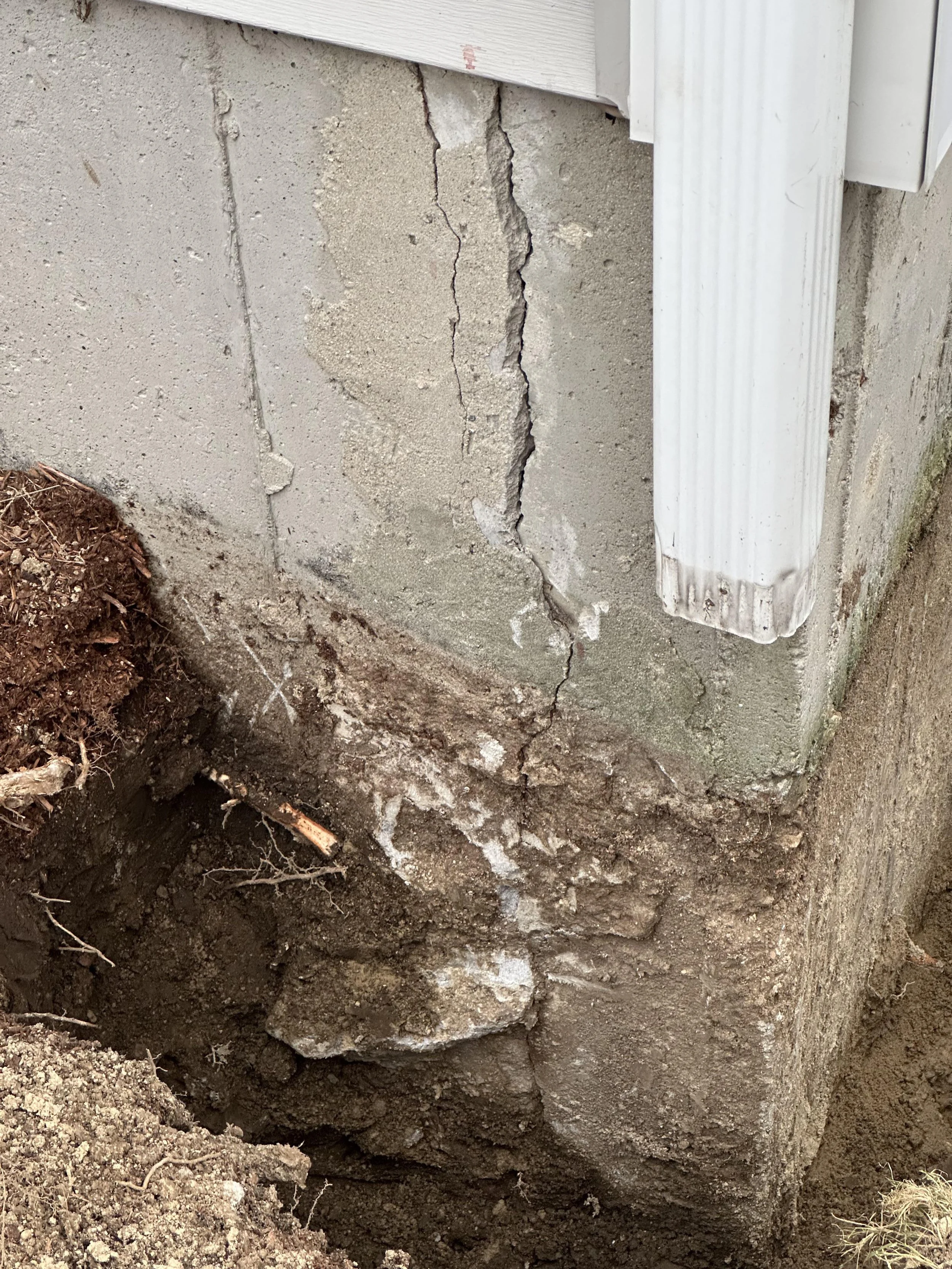 Close-up of a cracked concrete foundation wall with exposed soil and roots at the base, next to white siding and a section of a white downspout.