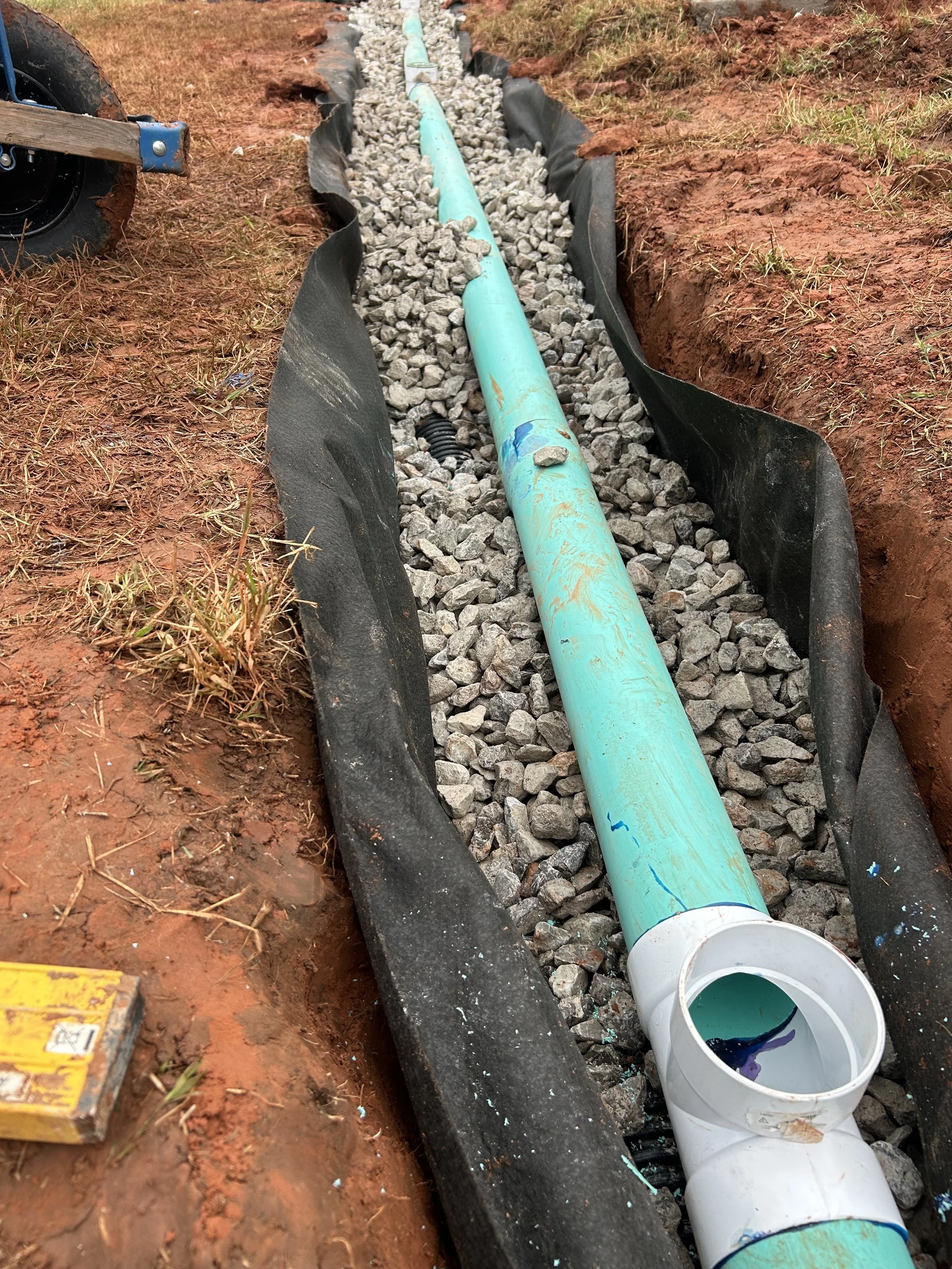 A construction site showing a new water pipe installation. A large turquoise pipe runs through a trench filled with gray gravel, lined with black plastic, with a white PVC pipe fitting at the end, and a bucket filled with purple dye nearby.