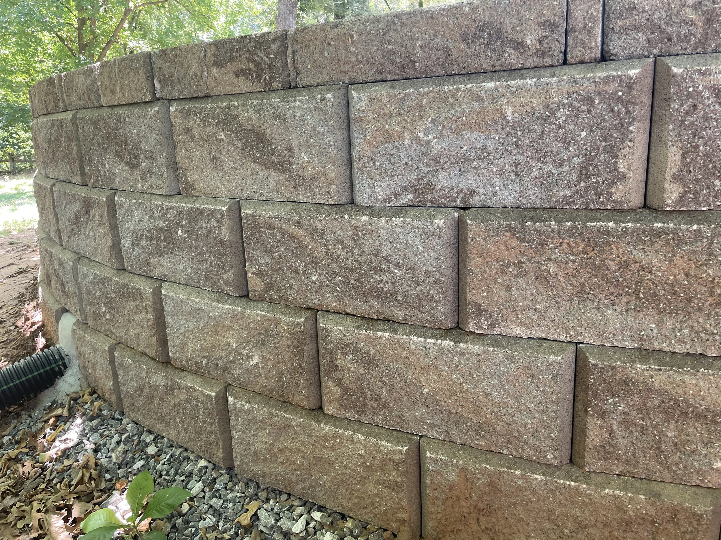Close-up of a stacked stone wall with large rectangular reddish-brown bricks, surrounded by trees and foliage, with leaves on the ground and a bendable black drainage pipe at the bottom left.