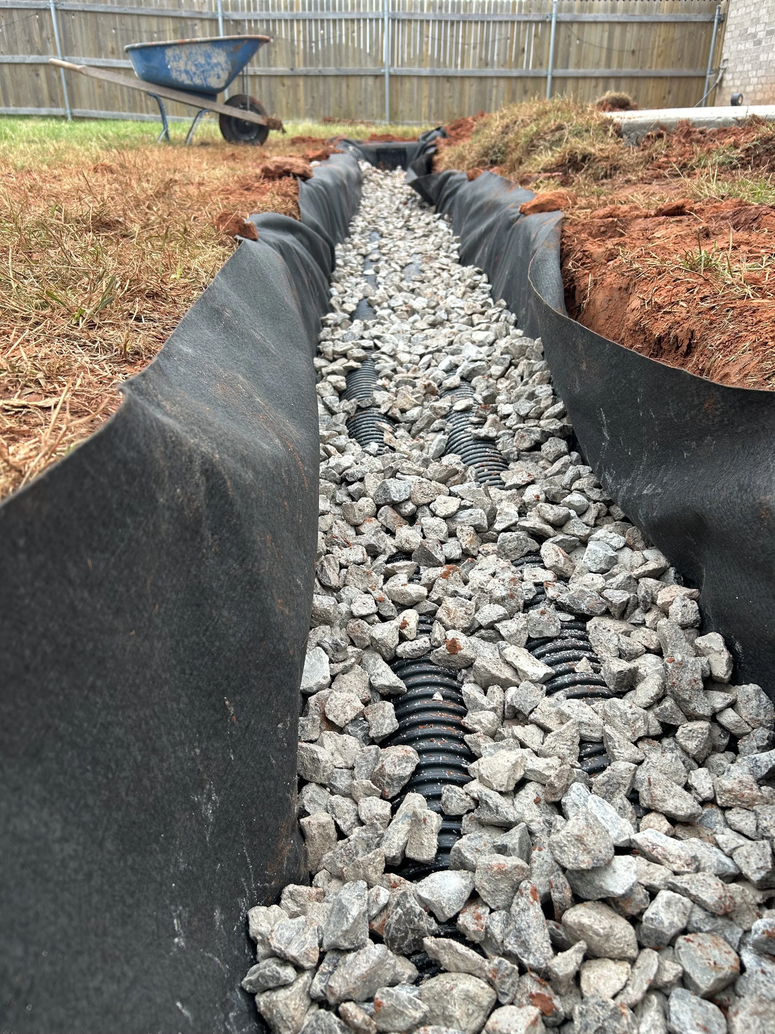 A trench filled with a geogrid fabric, gravel, and black tubing, with a wheelbarrow nearby and a wooden fence in the background.