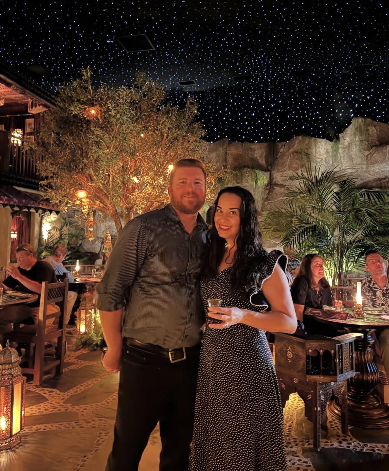 Sarah and Josh standing together in a restaurant with warm lighting, surrounded by tropical plants, a decorative tree, and people dining at tables in the background, under a dark ceiling with star-like lights.