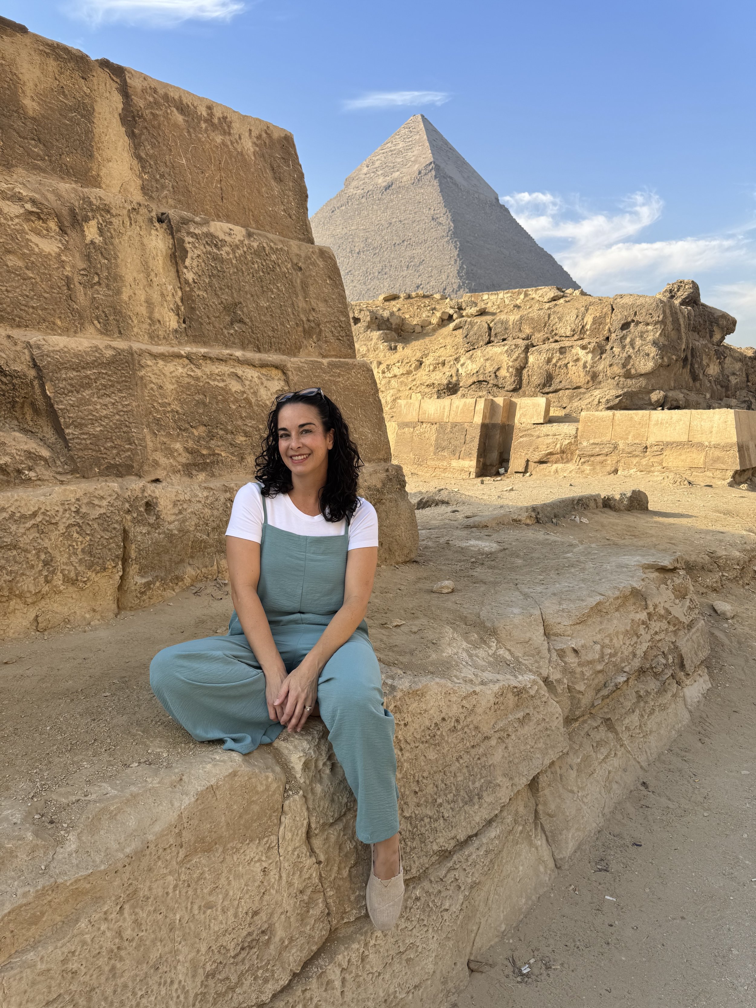 Sarah Durand Lathers sits on the ancient stone ruins of a pyramid in Egypt, with a larger pyramid visible in the background under a clear blue sky.