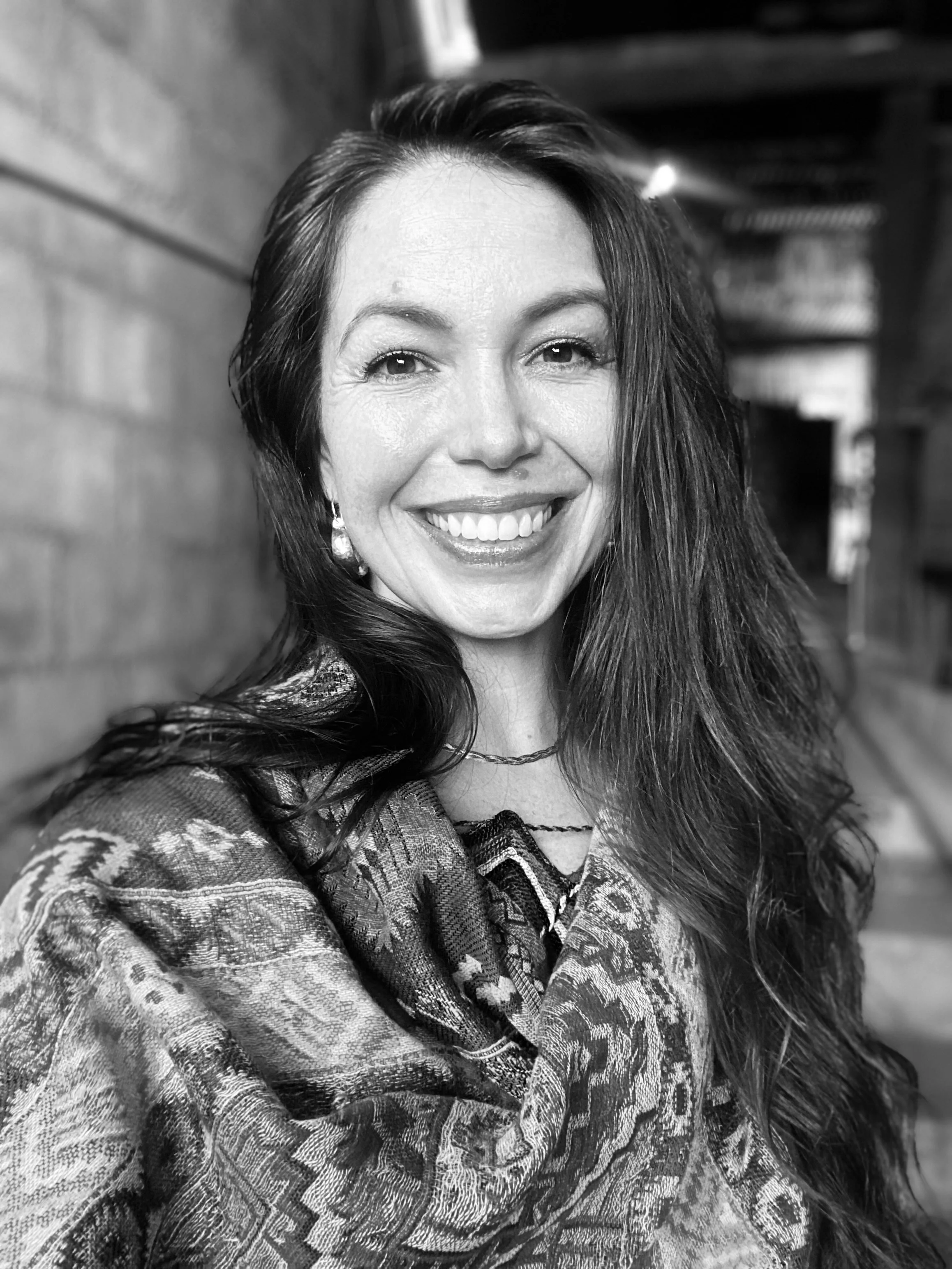A photo of Jessica Bell with long dark hair, earrings, and a patterned top, standing indoors near a brick wall.