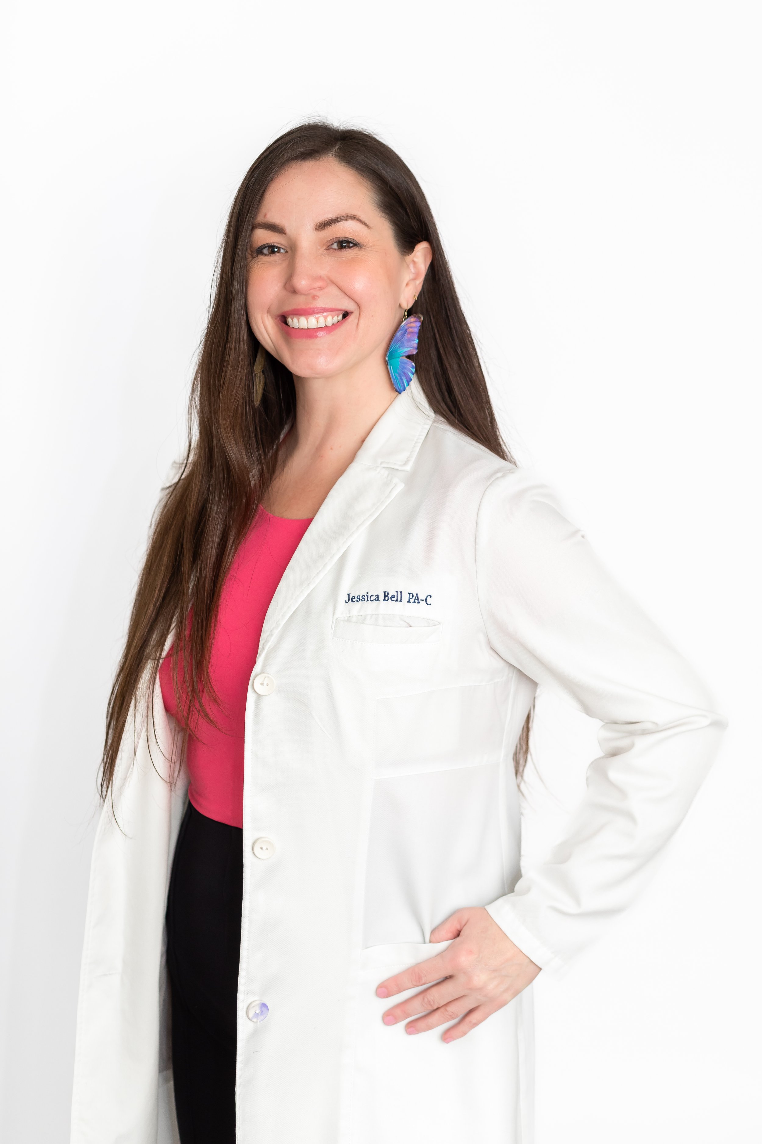 Smiling woman in a white lab coat with embroidered name Jessica Bell PA-C and colorful butterfly earrings, standing against a white background.