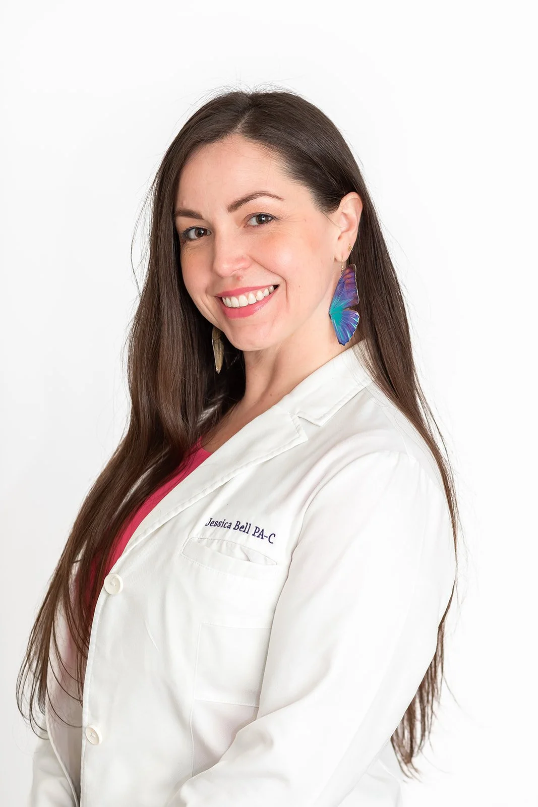 A smiling woman with long brown hair wearing a white medical coat and colorful butterfly earrings.