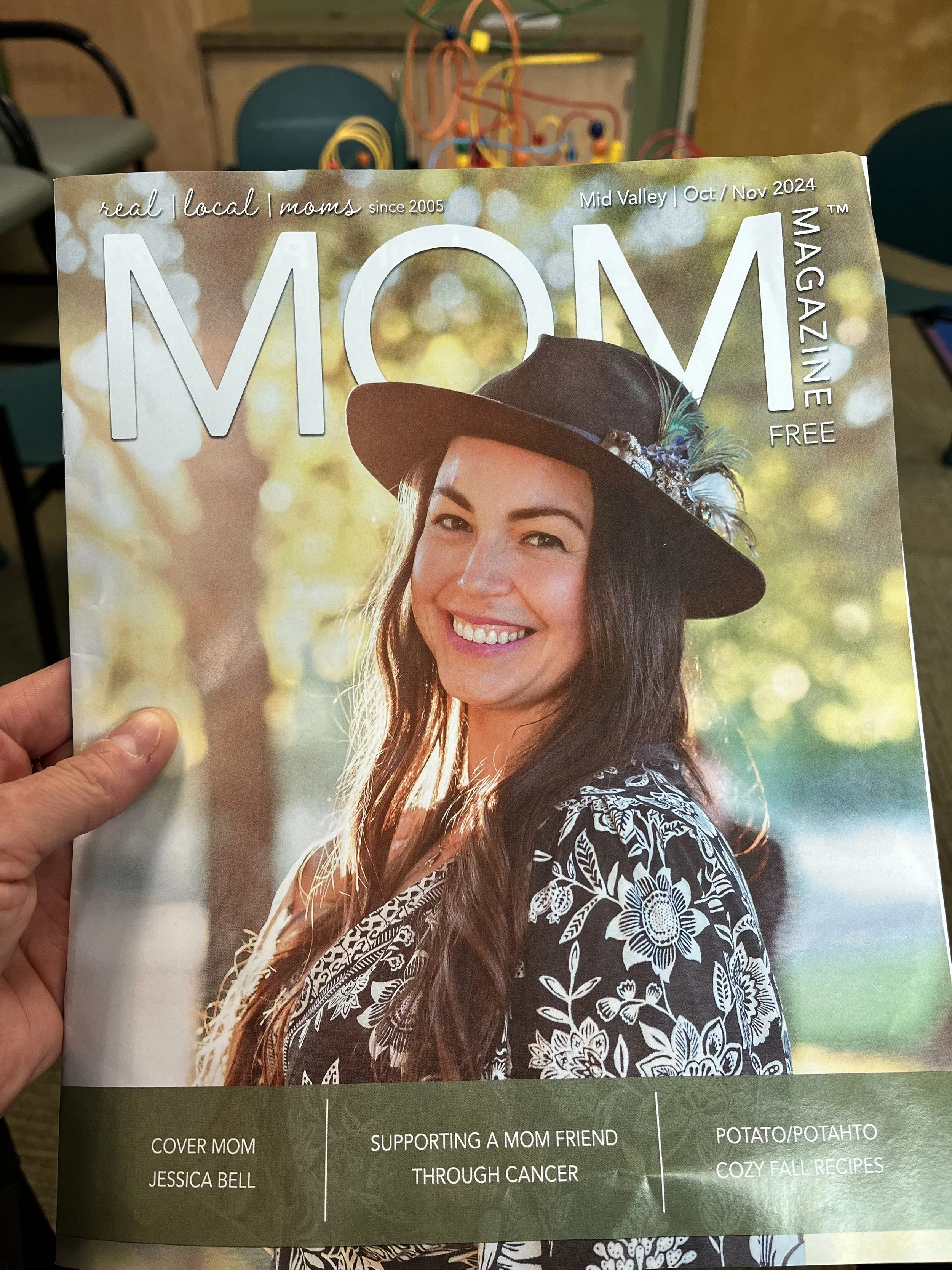 Cover of MOM magazine featuring Jessica Bell wearing a brown hat with feathers, and a black and white floral dress, outdoors with sunlight filtering through trees in the background.