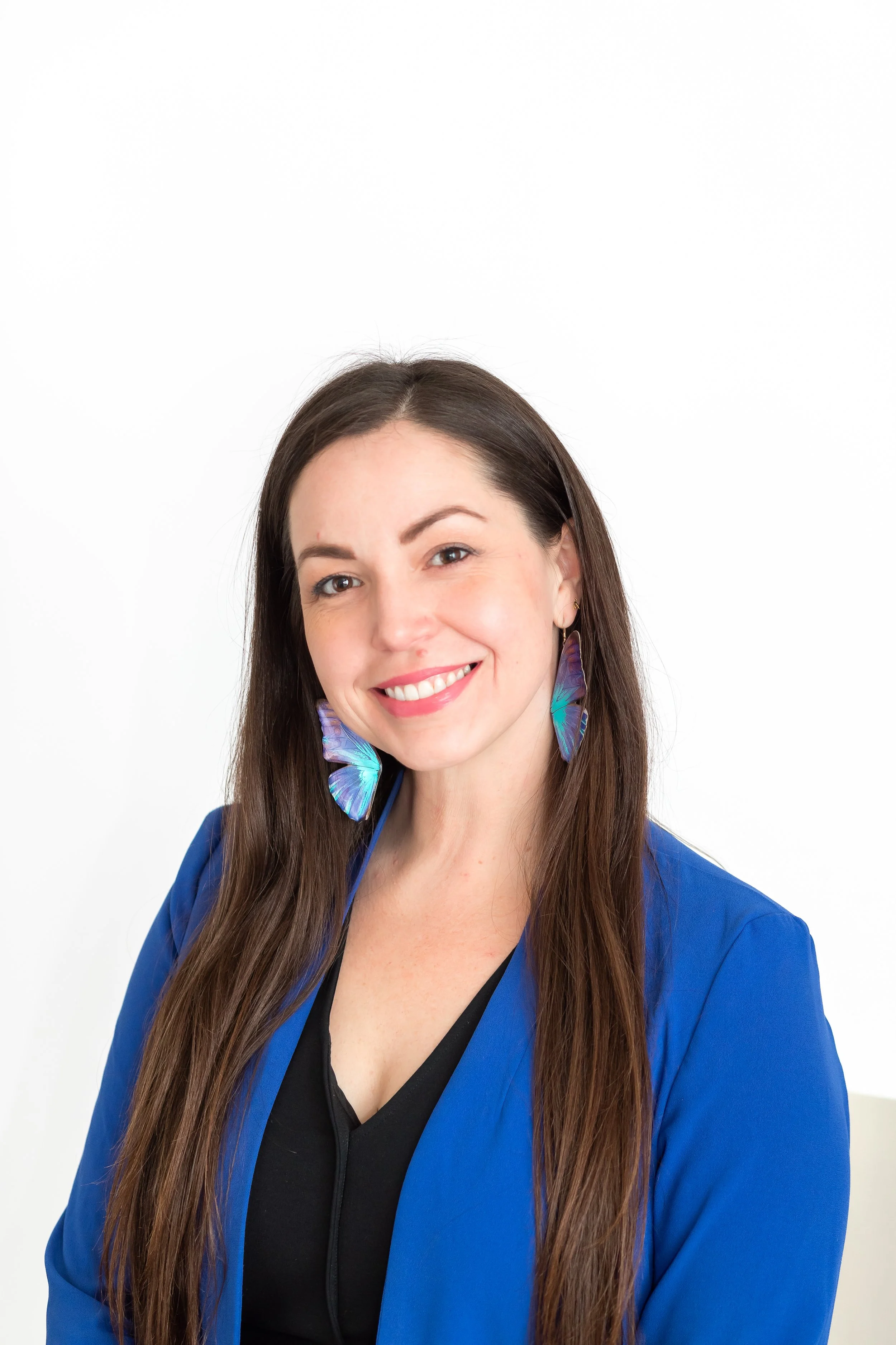 A woman with long brown hair and blue eyes, smiling, wearing large blue earrings resembling butterflies, a blue blazer over a black top, posed against a plain white background.