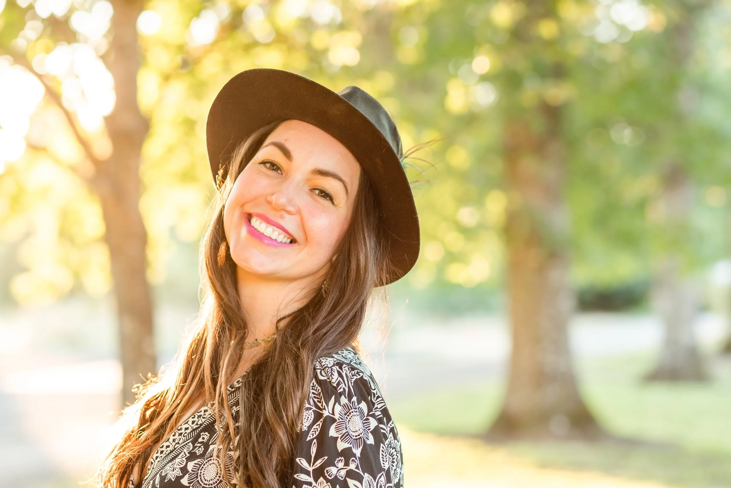 A photo of Jessica Bell smiling outdoors in a park, wearing a brown wide-brimmed felt hat and a black and white floral patterned top, with trees and sunlight in the background.
