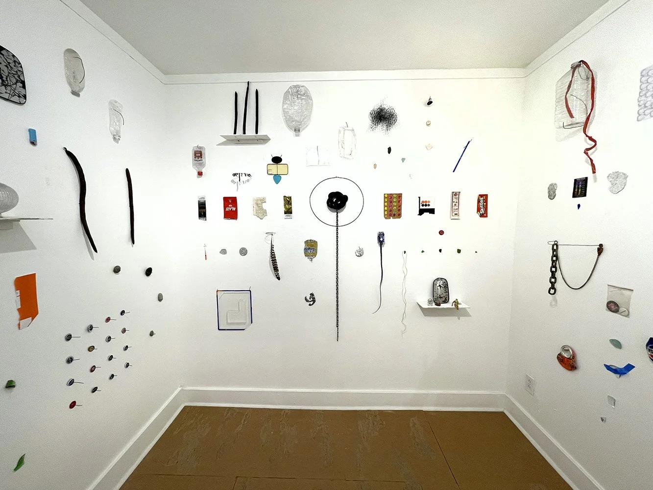 An art installation on a white wall featuring various small objects and materials found on the street near the gallery arranged in a collage style, with some objects hanging or attached to the wall.