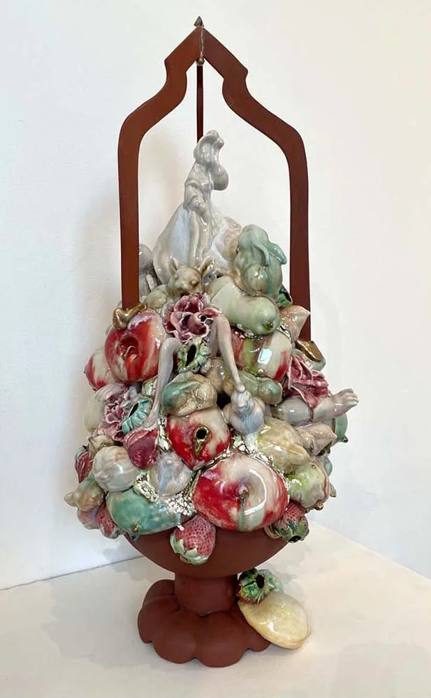 An artistic sculpture consisting of various ceramic fruit and vegetable elements, with some human and animal figures, arranged inside a wooden frame that resembles a hanging bath or vessel.