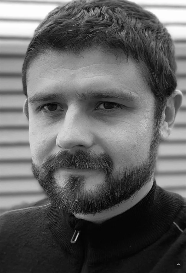 A black and white close-up of a man with short dark hair, a beard, and a mustache, wearing a dark zip-up sweater, standing in front of a horizontal slatted background.