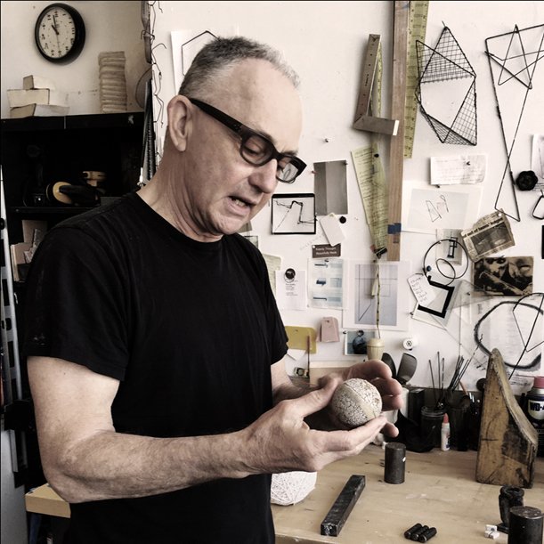 A man with glasses and a black T-shirt examining a stone in his hands in a workshop filled with tools and sketches on the wall.