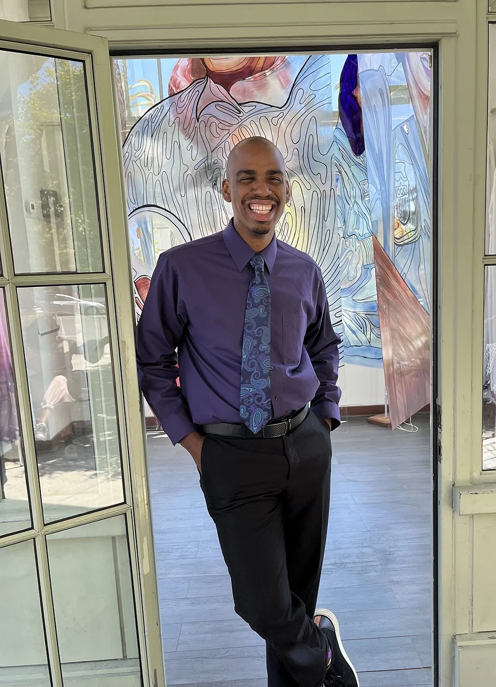 tall man with bald head and mustache wearing a shirt and tie standing in a doorway with painted plexiglass panels behind him
