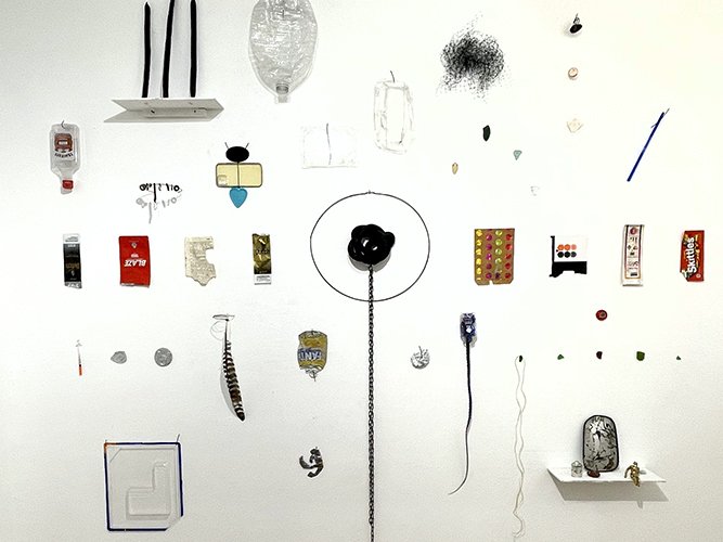A white wall decorated with various small objects  found on the street near the gallery,including a black chain, a small shelf with colored objects,.