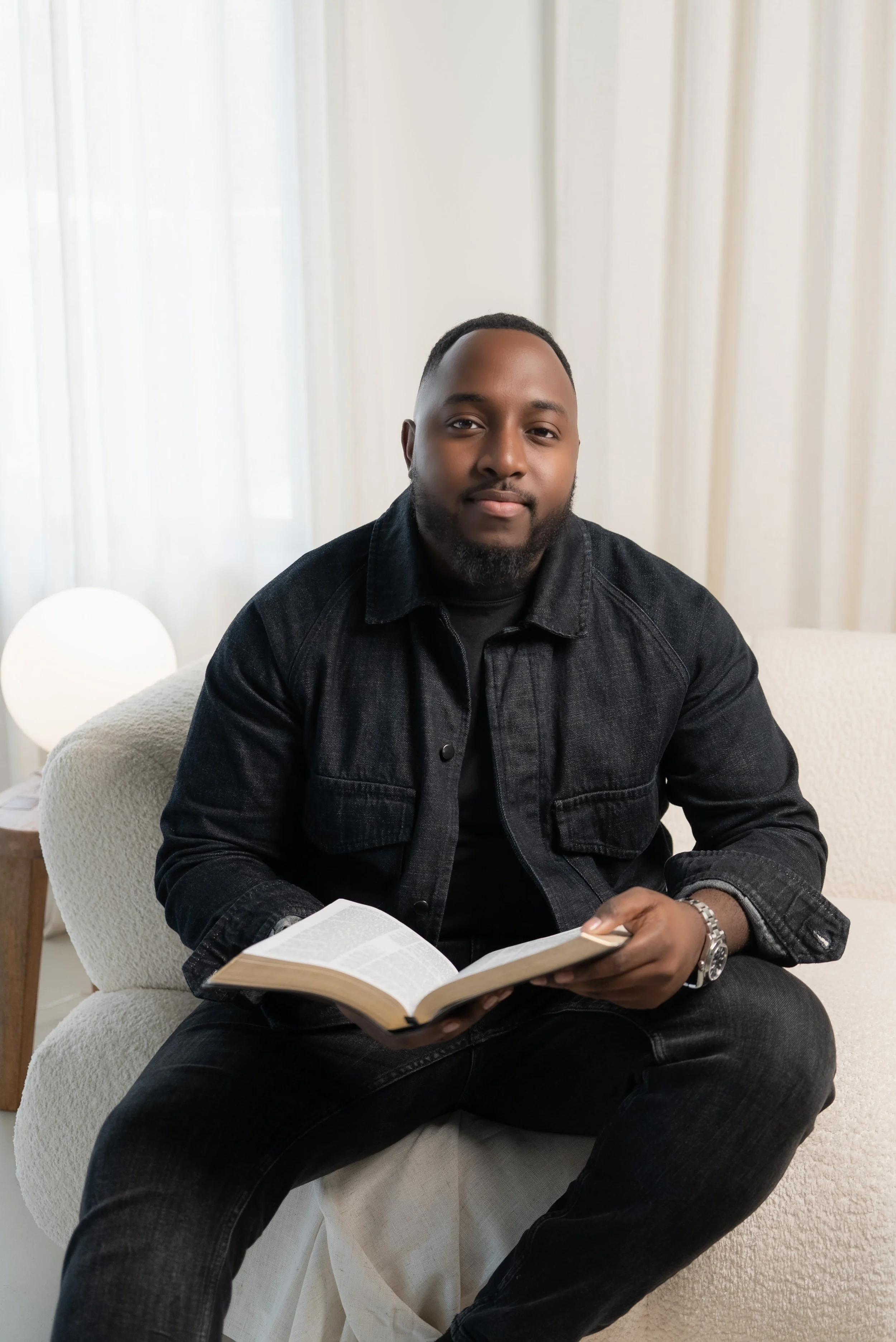 A man is sitting on a beige couch, holding an open book, with a calm expression. He is wearing a black denim jacket and black jeans, and has a watch on his left wrist. The background features white curtains and a round white lamp.