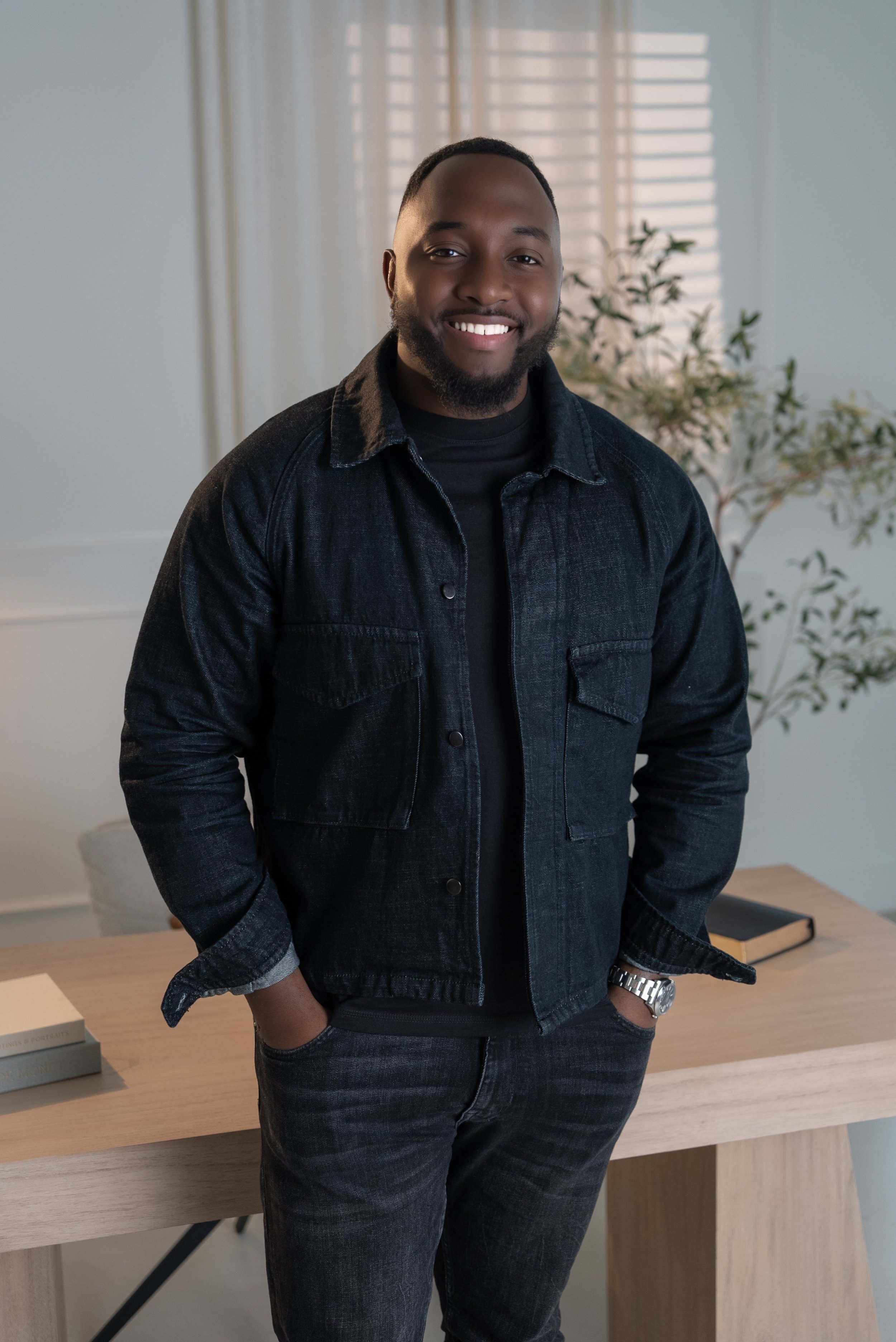 A smiling man with a beard wearing a dark denim jacket and black shirt, standing with hands in pockets in a modern, well-lit room with a wooden table, books, and a potted plant in the background.