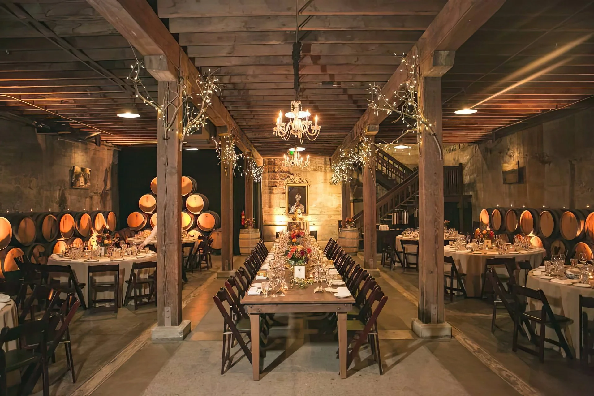 A rustic banquet hall decorated with fairy lights, chandeliers, and wine barrels, set for a formal dinner with round and rectangular tables, floral centerpieces, and elegant place settings.