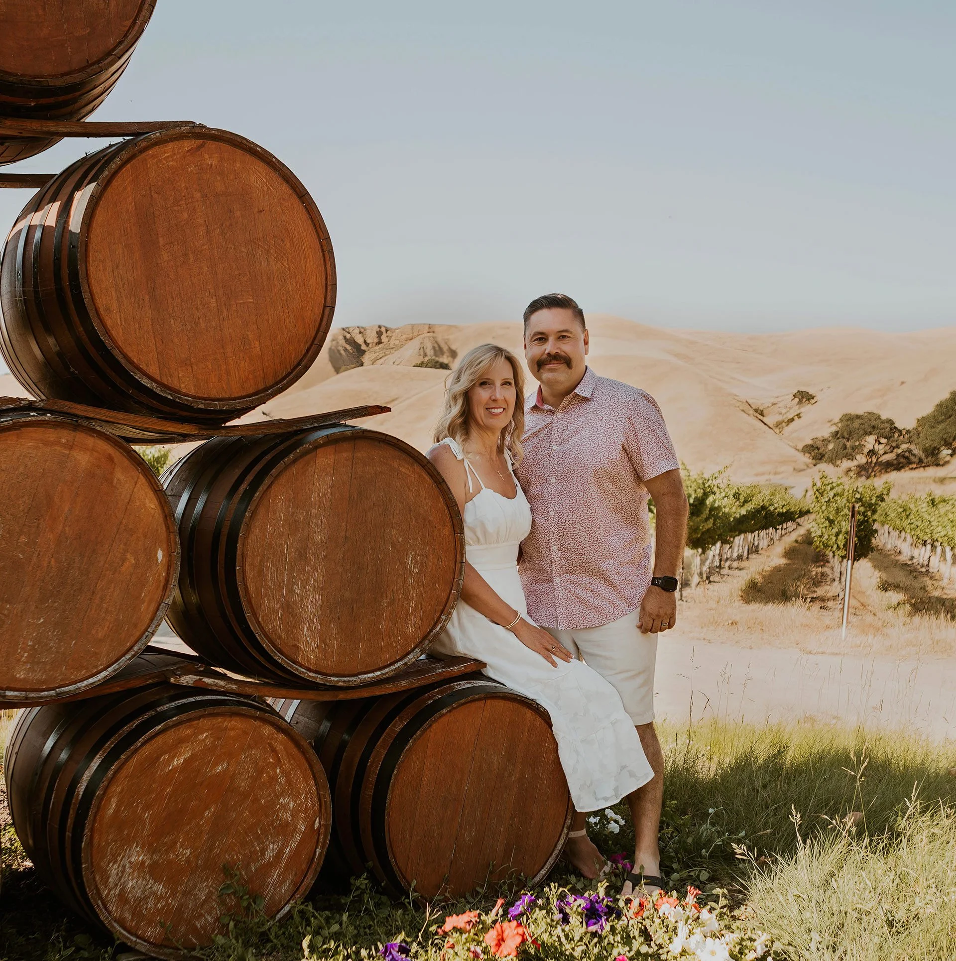 A couple standing next to a stack of wine barrels in a vineyard with rolling hills and grapevines in the background.