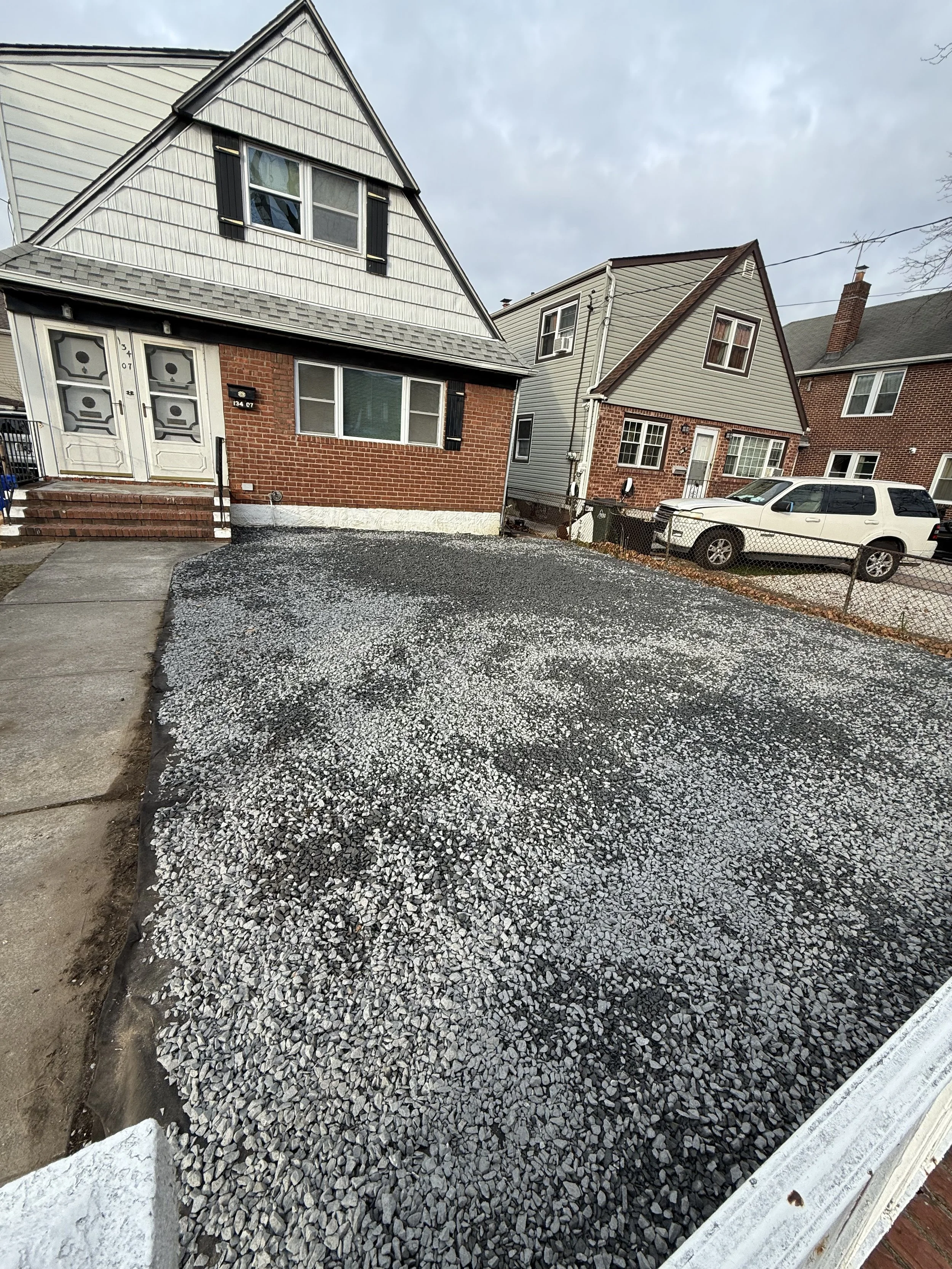A gravel driveway in front of a house with a brick and siding exterior, with neighboring houses and parked cars visible.