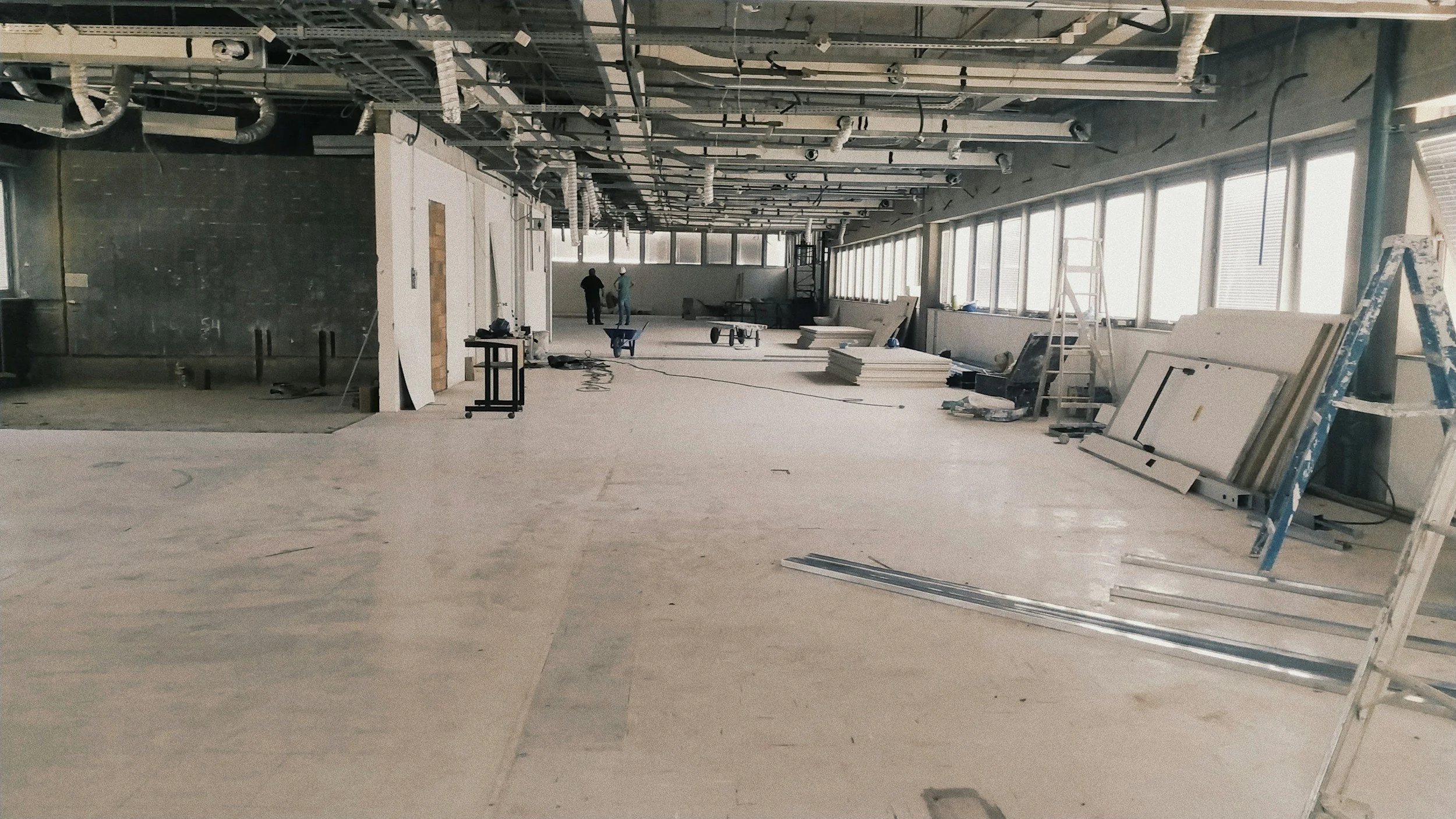 Interior of a building under construction with workers, ladders, construction materials, and equipment scattered around, and large windows along the right side.
